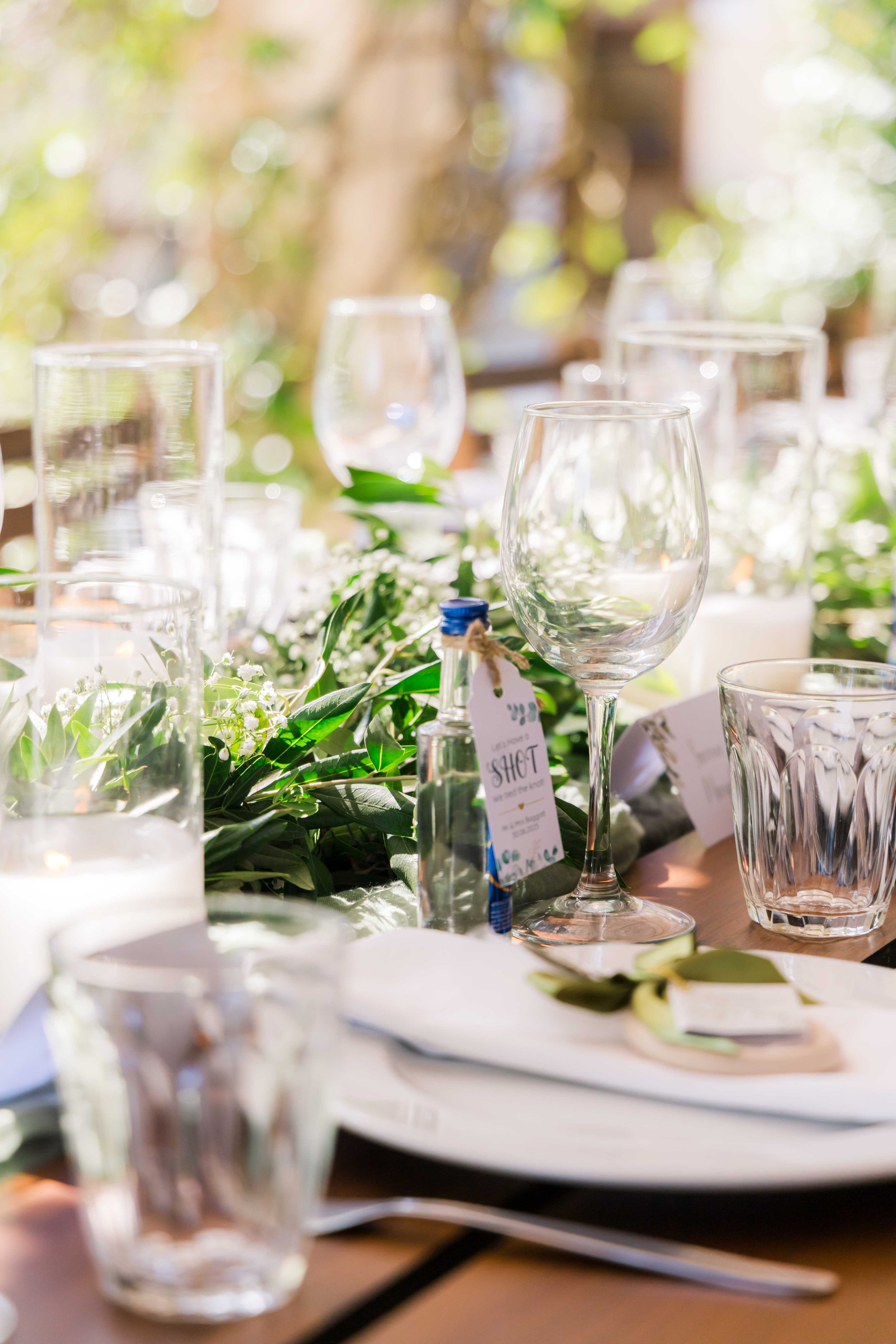 Elegant outdoor table setting with glassware, white flowers, greenery, and place cards, illuminated by natural sunlight.