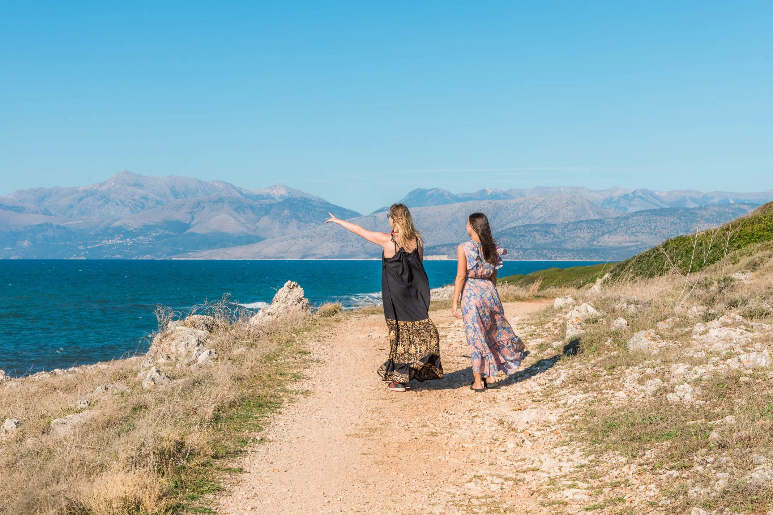 Two women walk along a rocky, dirt path beside the ocean with mountains in the background. One woman points toward the mountains while they enjoy a sunny day.