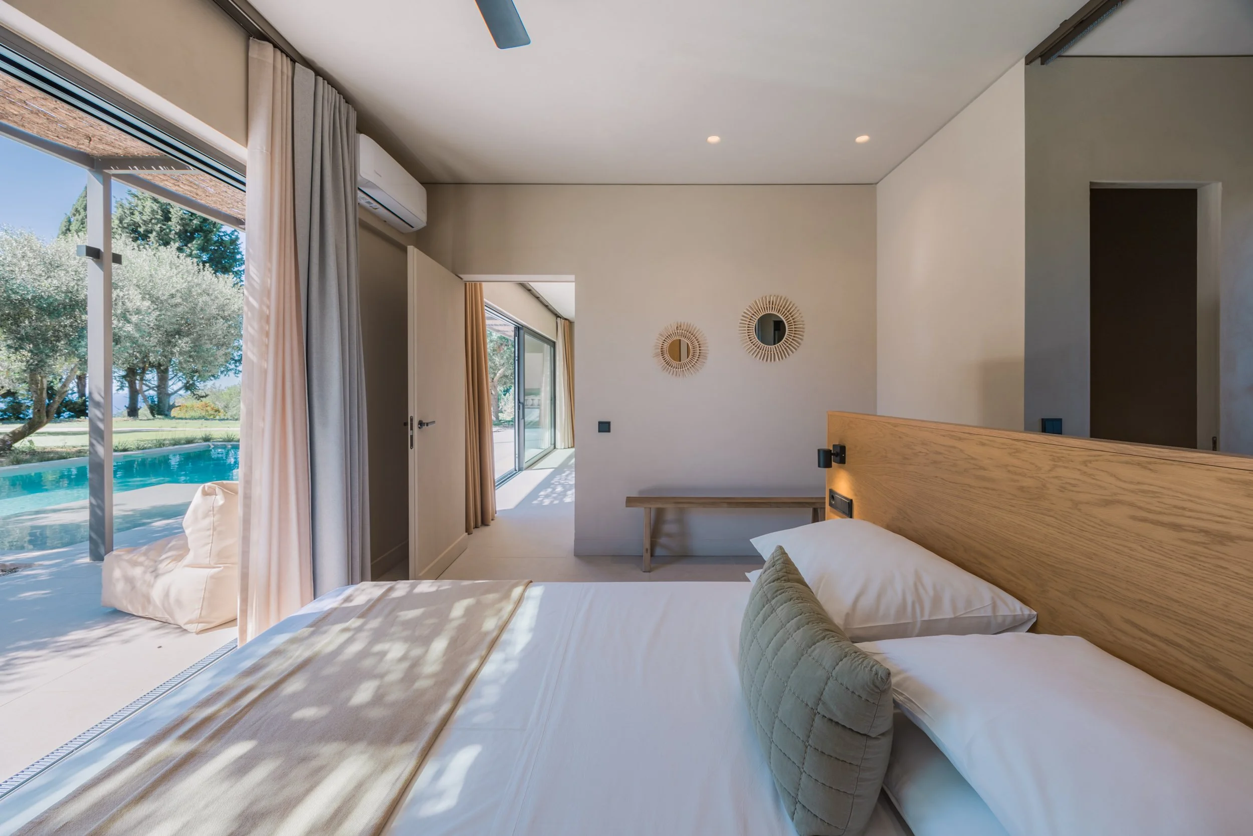 Modern bedroom with large sliding glass door leading to outdoor pool and garden, featuring a wooden headboard, white bedding, and beige curtains.