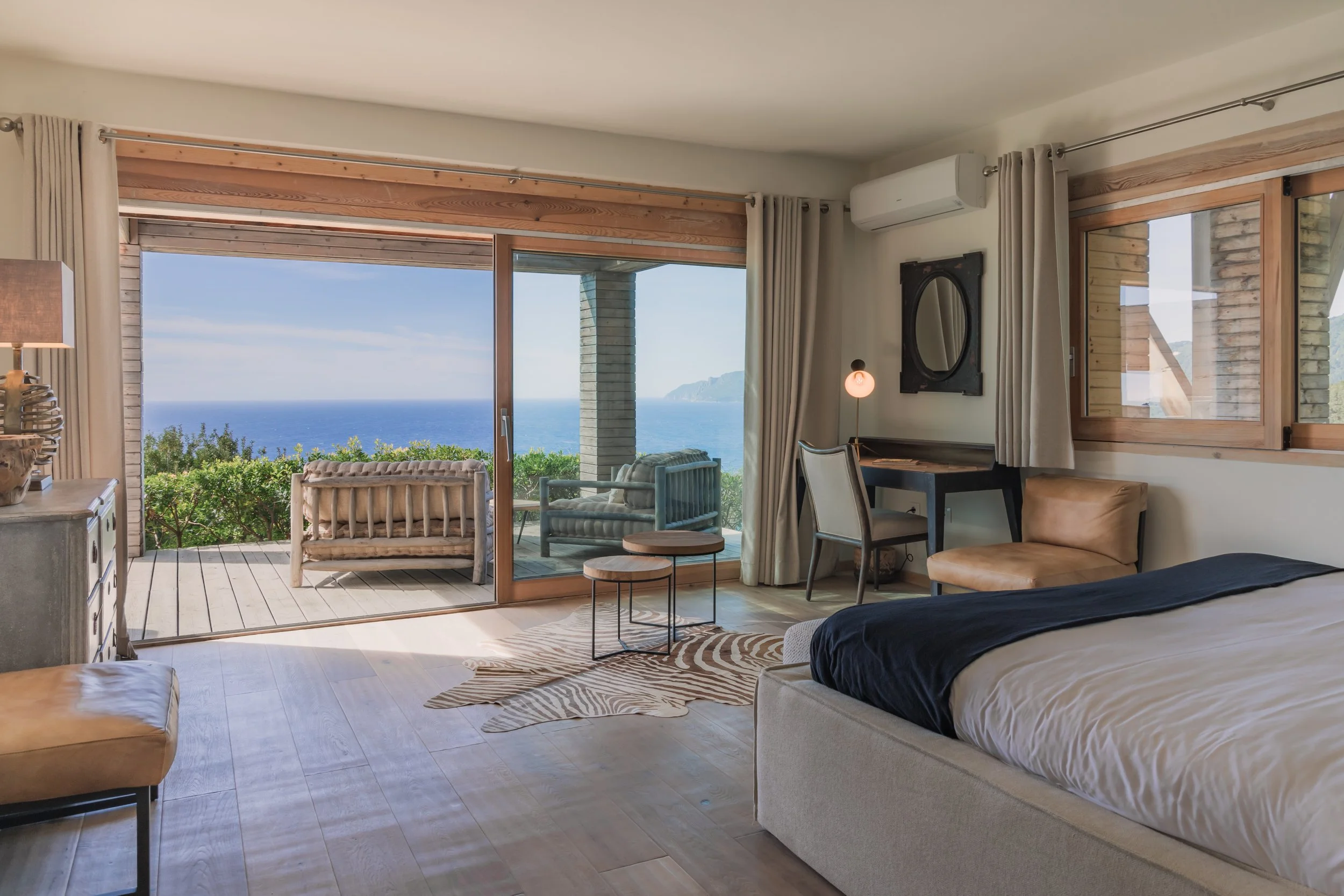 A bedroom with a sliding glass door leading to a balcony overlooking the ocean, with outdoor seating and greenery.