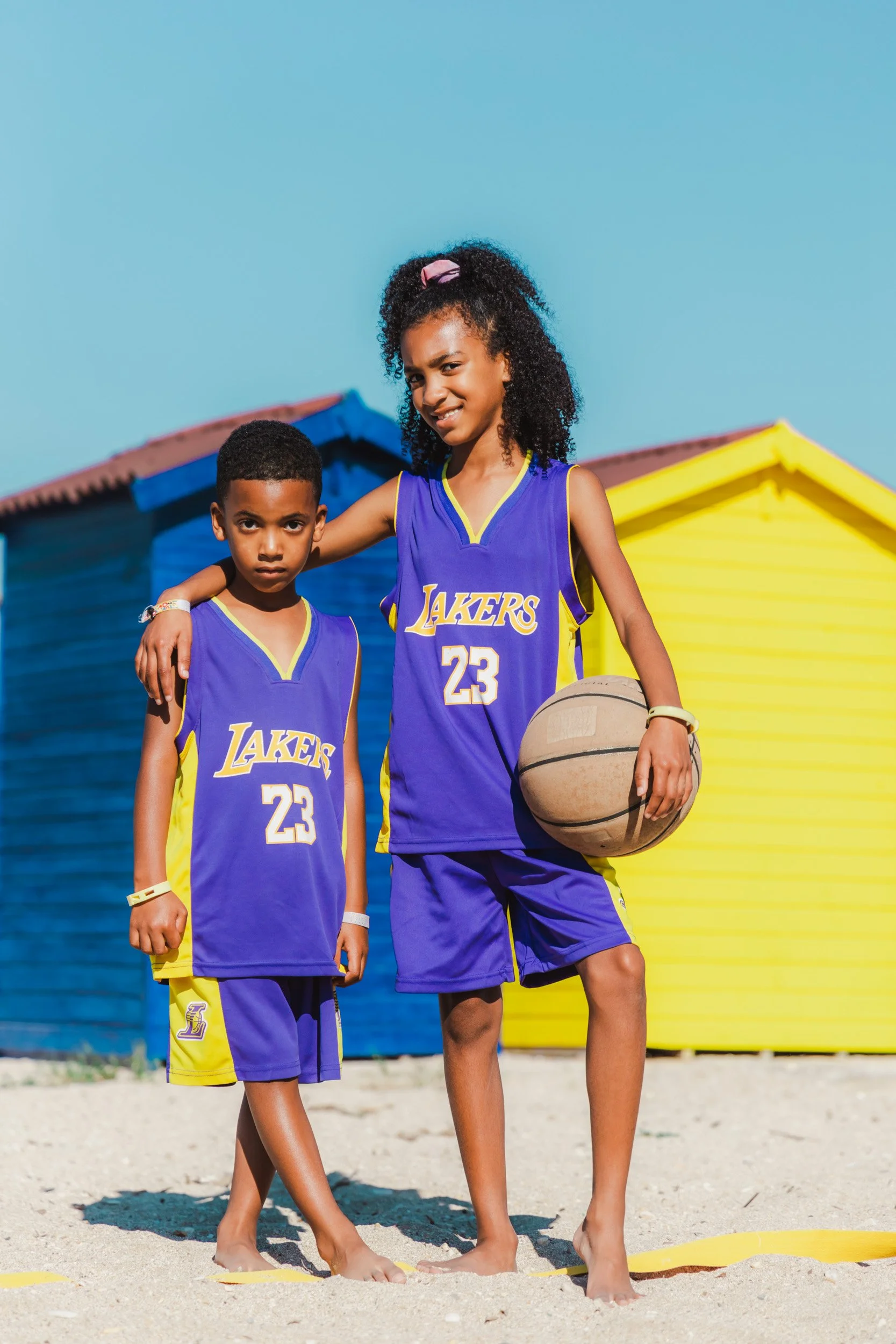 Two children in Los Angeles Lakers basketball uniforms standing on a sandy beach with colorful beach huts in the background. The older girl has her arm around the younger boy, and she is holding a basketball.