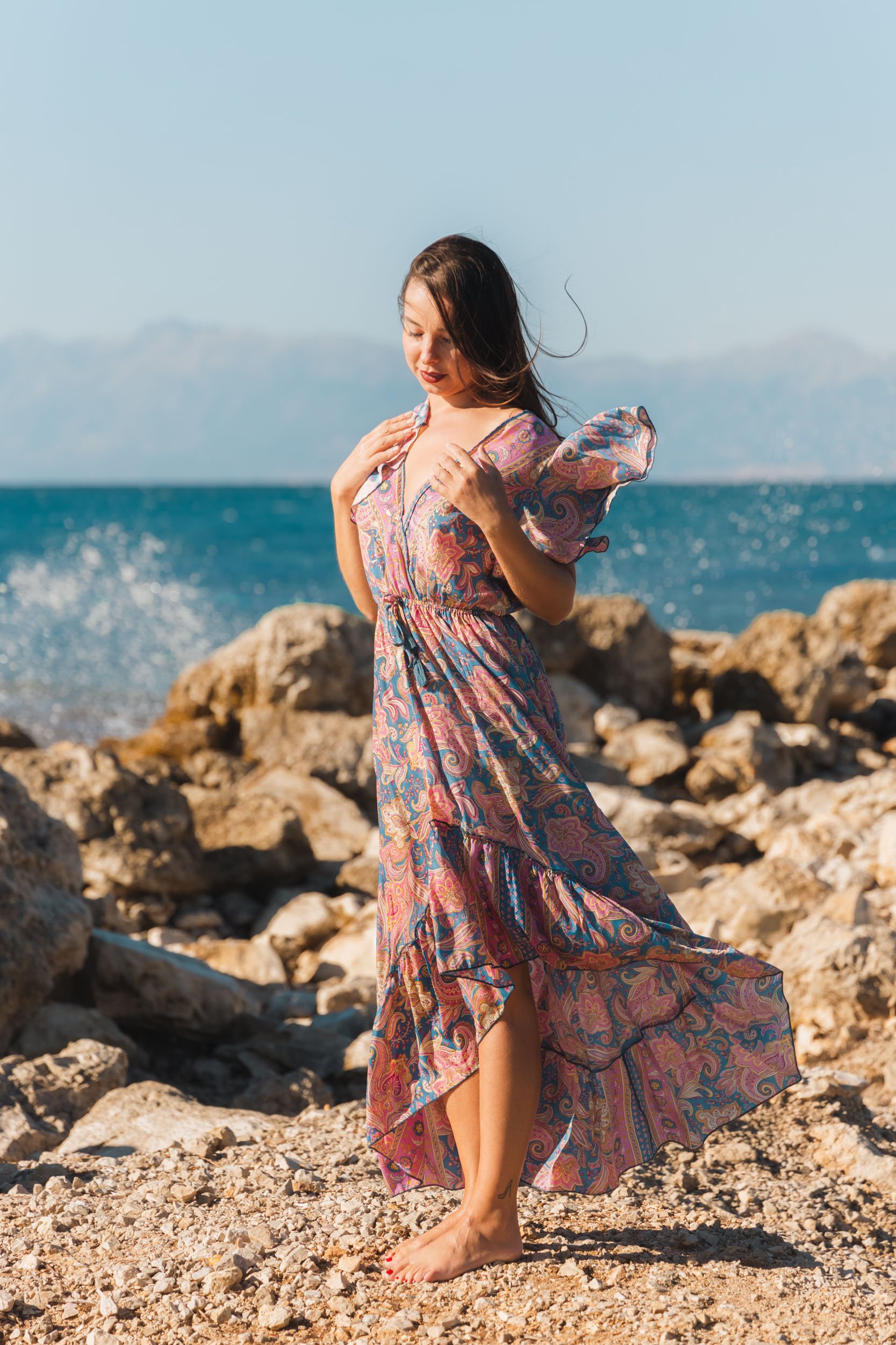 A woman standing barefoot on a rocky beach near the ocean, wearing a colorful, patterned maxi dress with puff sleeves, with the wind blowing her hair.