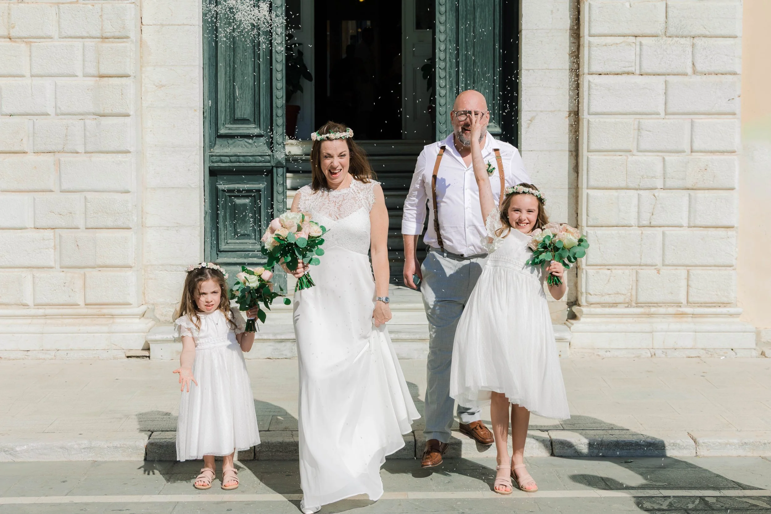 A wedding scene with four people, including a bride in a white dress holding a bouquet, a young girl in a white dress and floral headband, and a man and another young girl also holding bouquets, standing on steps in front of a building with open door