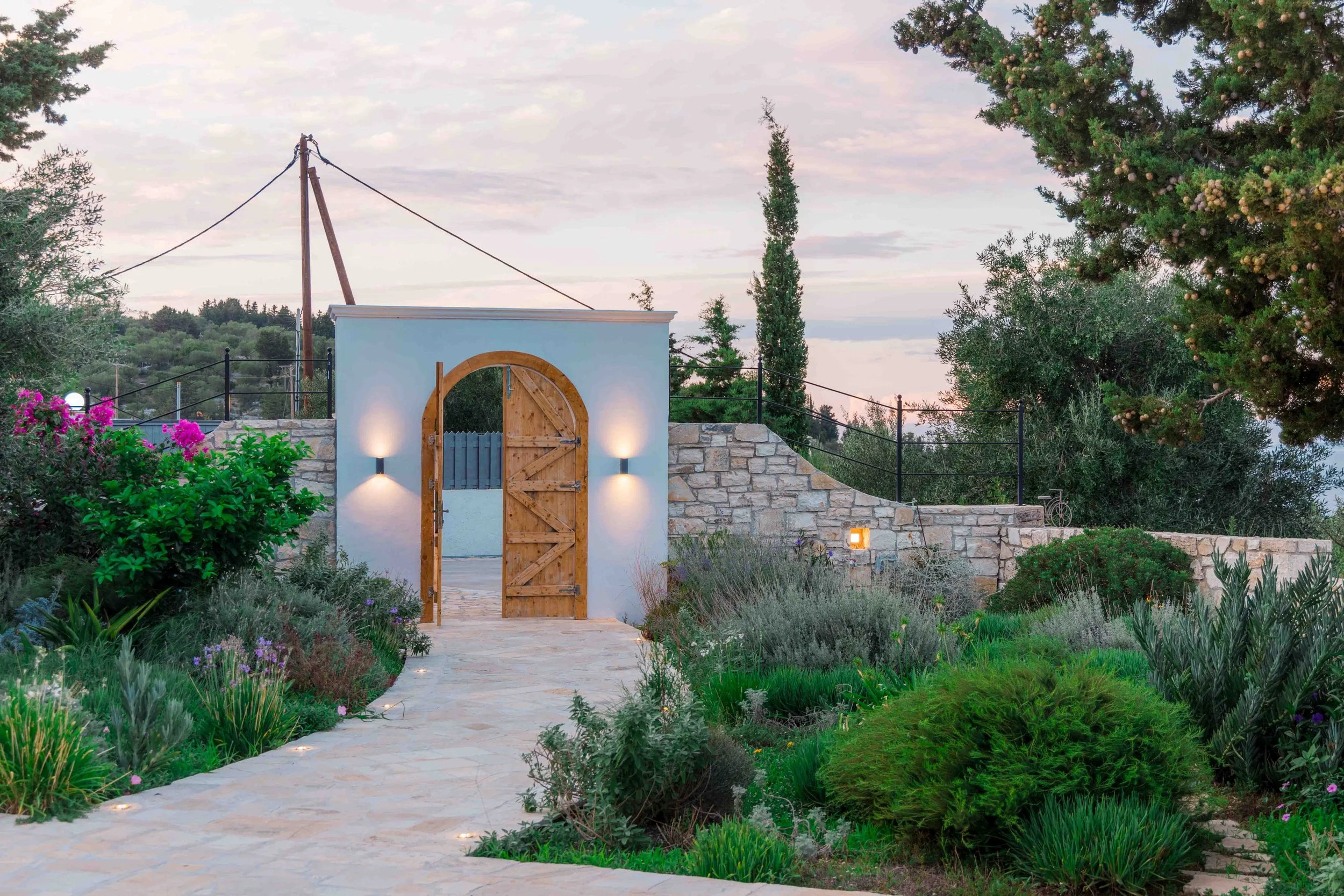 A picturesque garden pathway leading to a small white building with a wooden arched door and exterior lights. The garden features lush green plants and flowers, with a stone wall and black metal railing in the background. The sky is partly cloudy dur