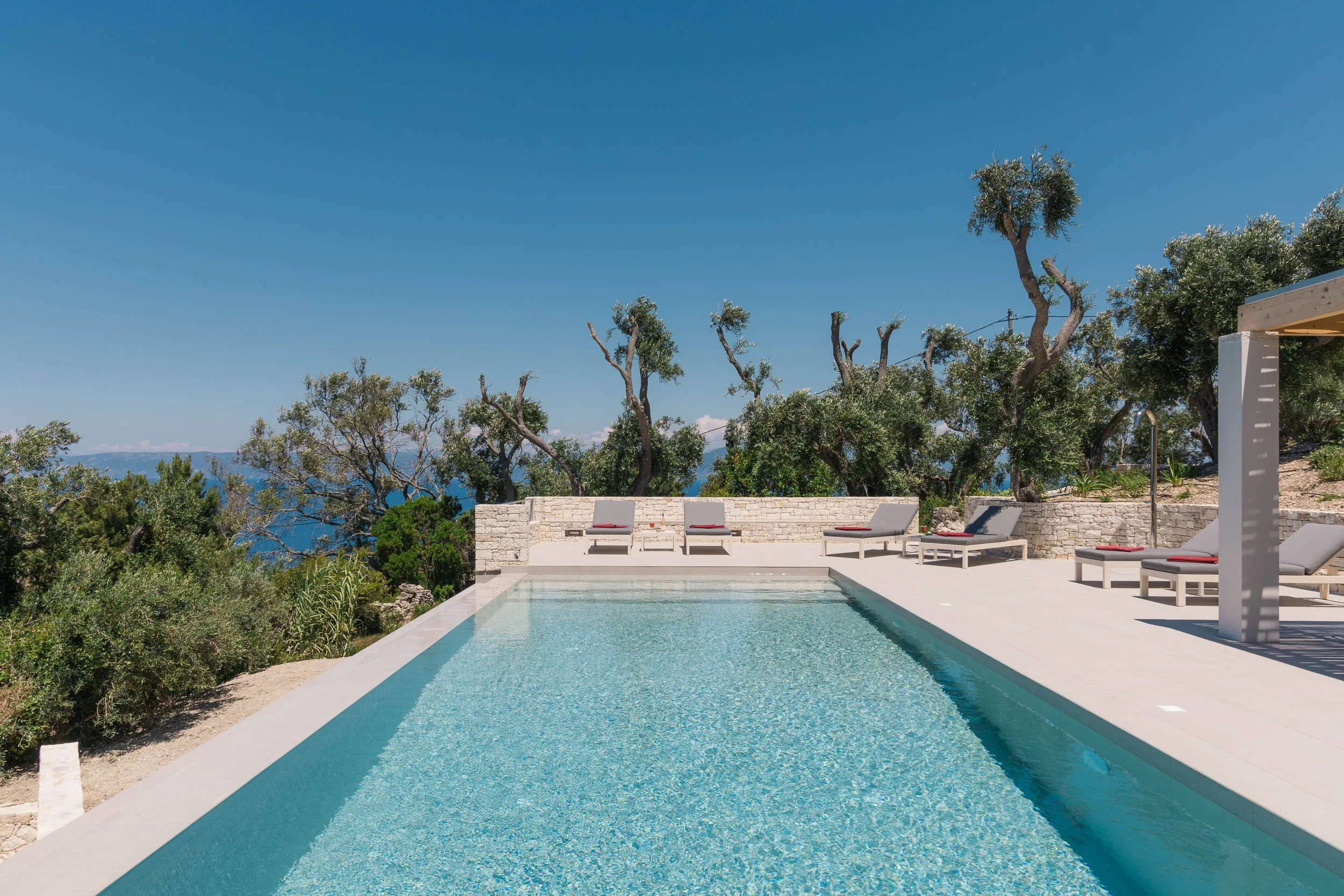 Swimming pool surrounded by gray lounge chairs with yellow cushions, set against a backdrop of trees and blue sky at a sunny day.