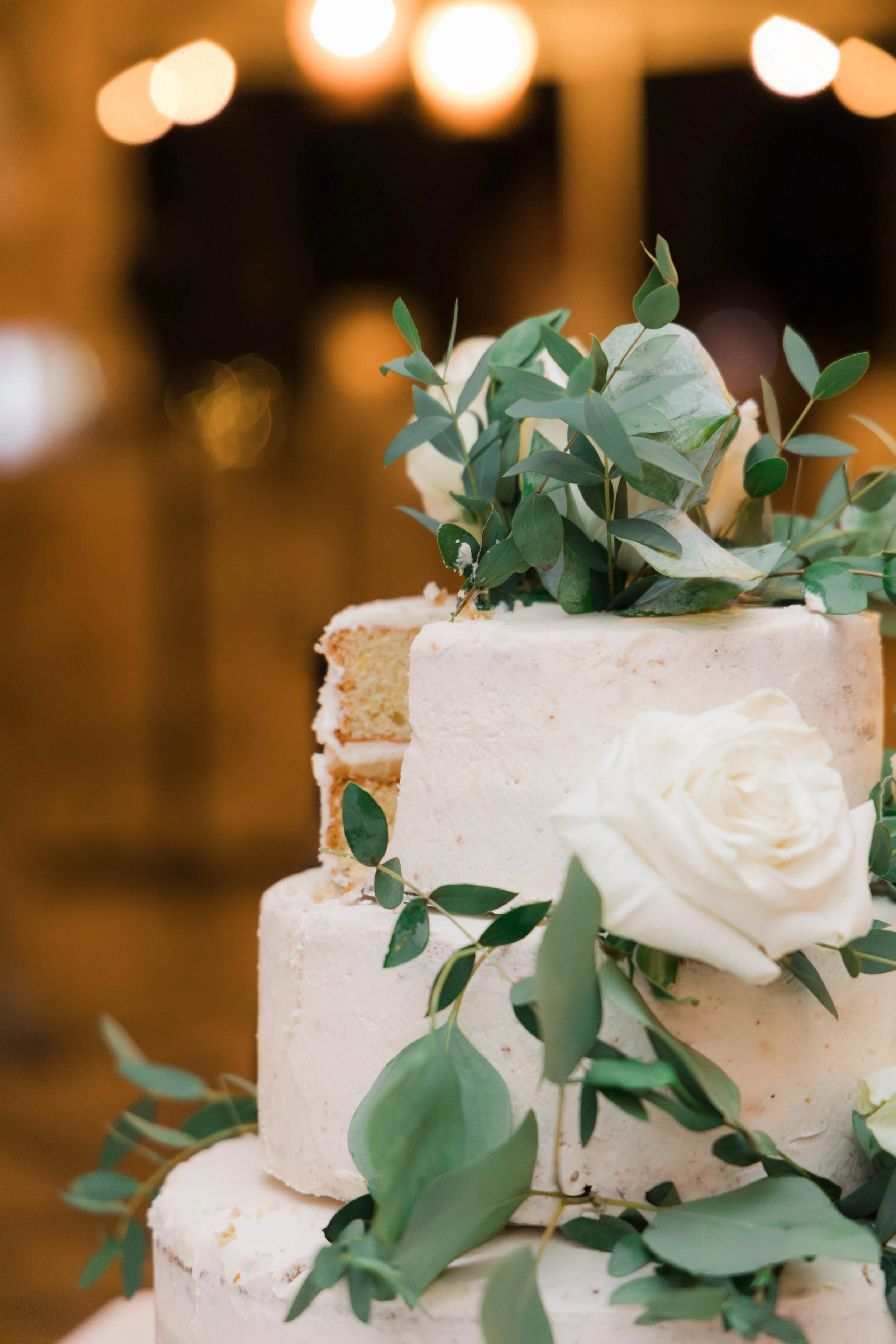 A close-up of a white wedding cake decorated with white roses and green leaves, with a blurred background featuring warm lighting.