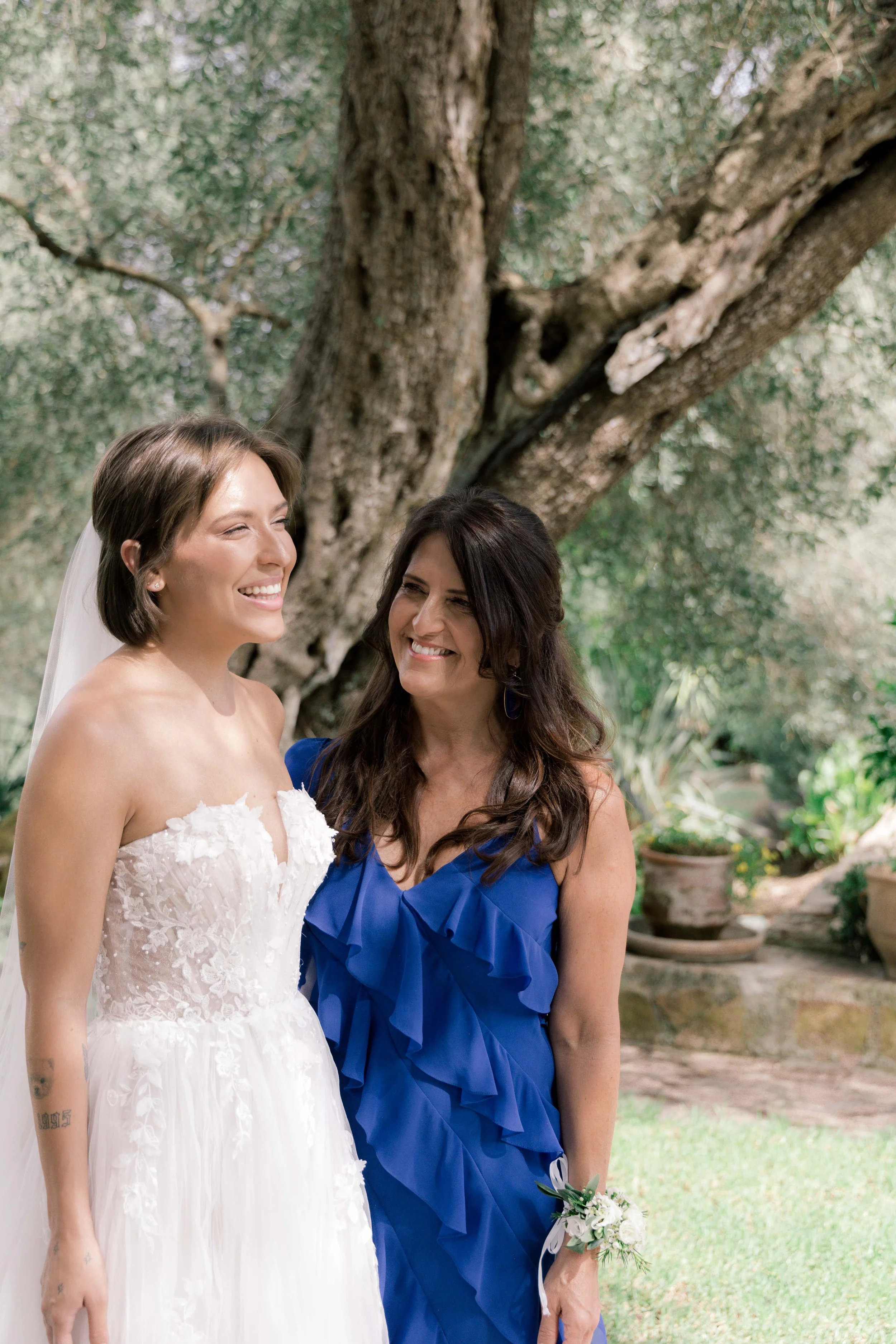 A bride in a wedding dress standing outdoors with a woman in a blue dress, both smiling, in front of a large tree.
