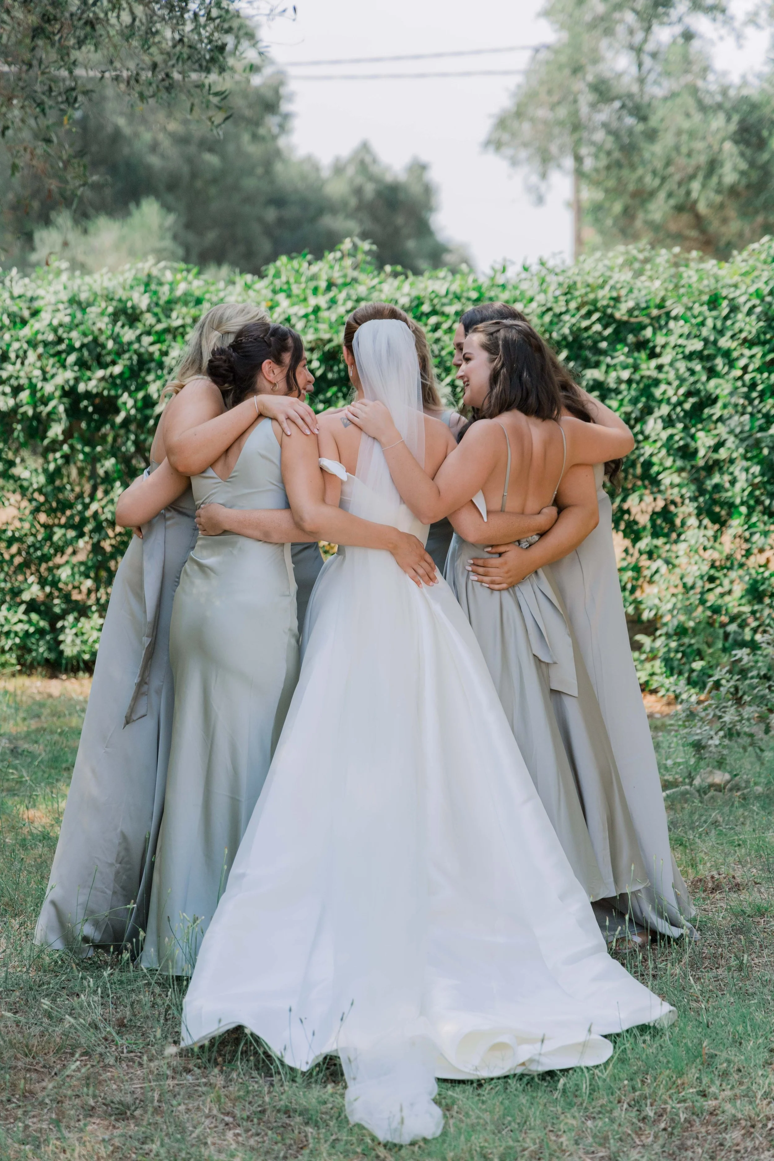 A group of women, including a bride in a white wedding gown and veil, hugging and smiling outdoors with green shrubbery in the background.