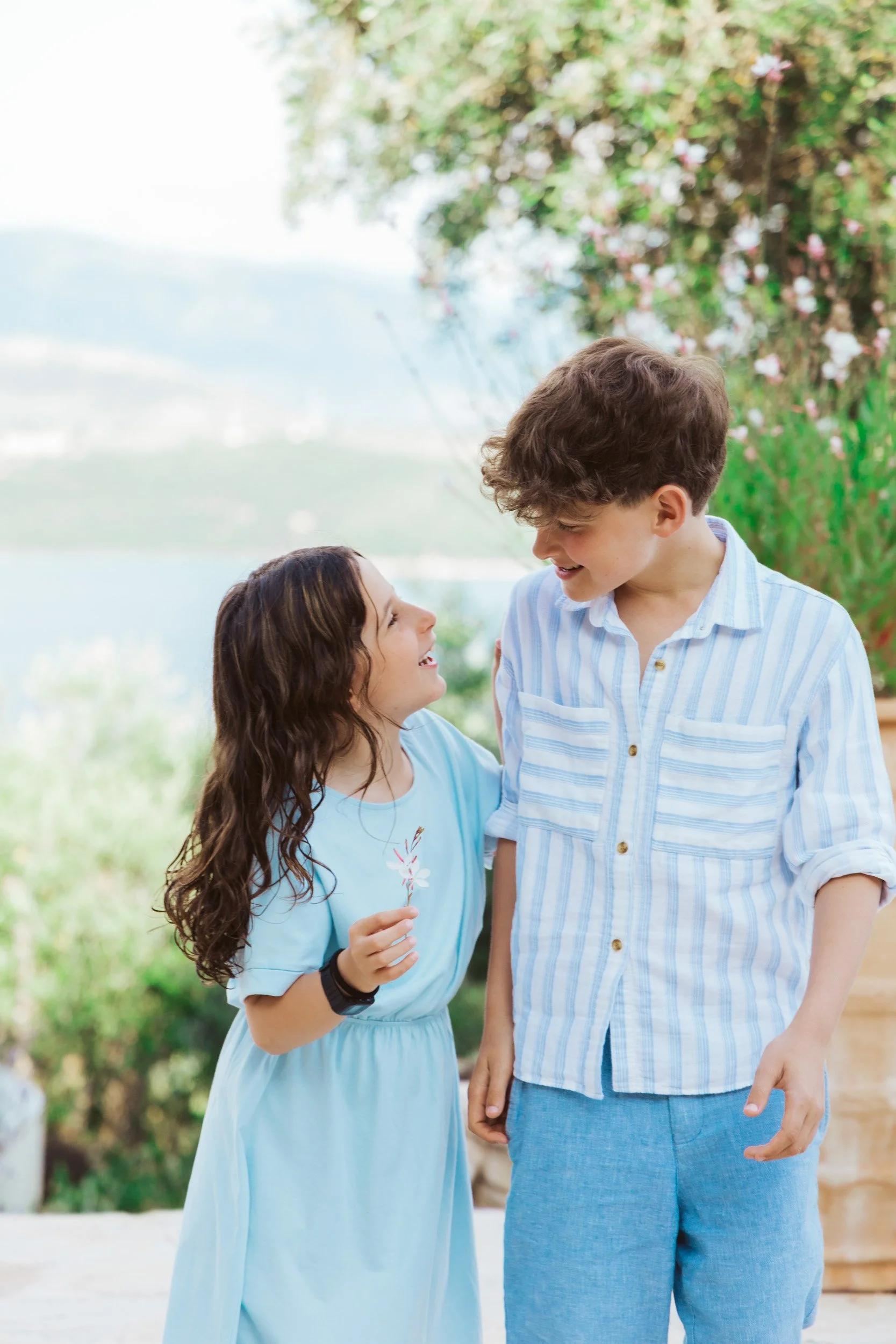 A young girl with long curly hair holding a flower, smiling at a teenage boy with short curly hair, outdoors with greenery and lake background.