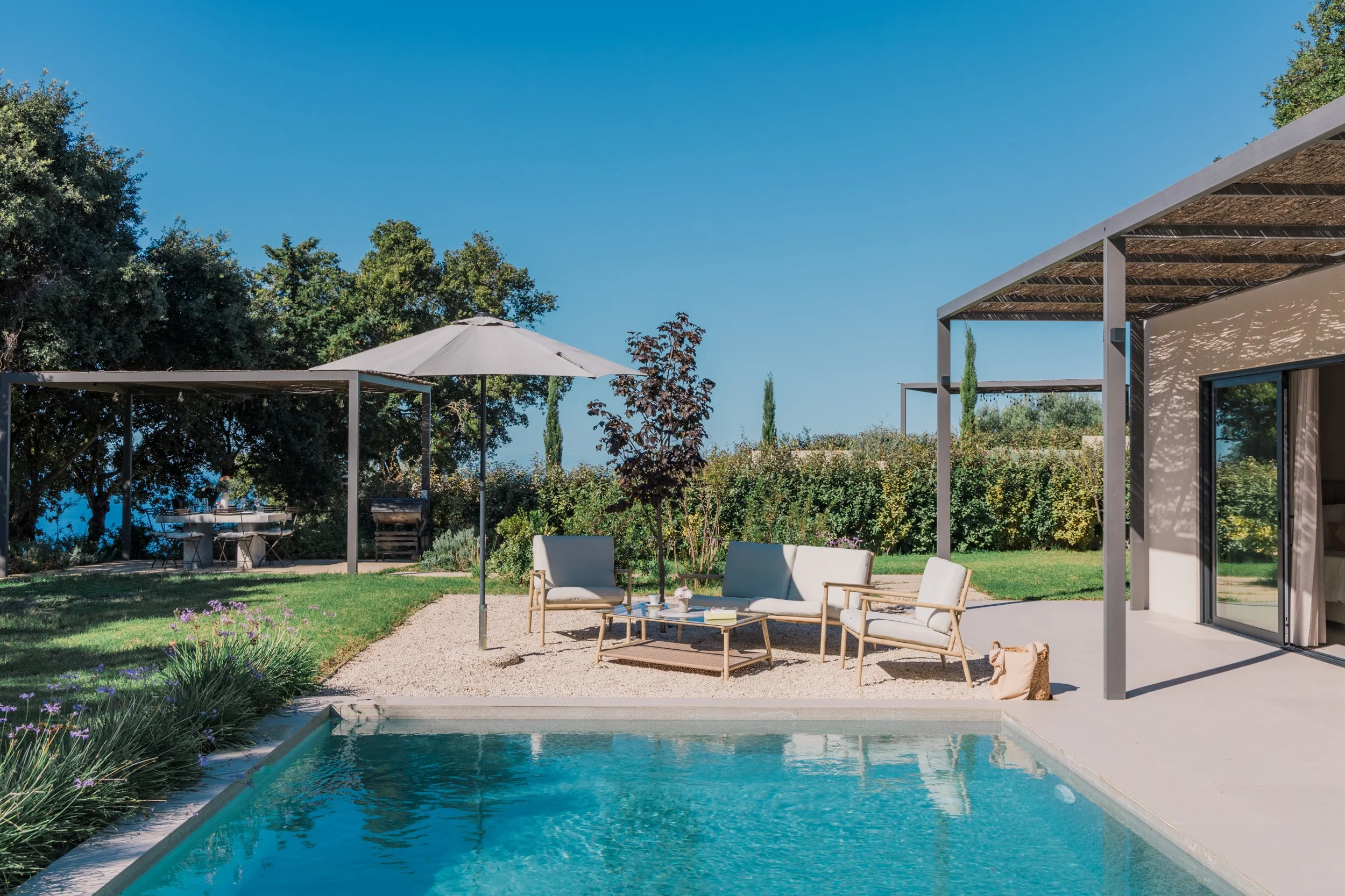 Modern backyard with a swimming pool, outdoor seating with white cushioned chairs and a sofa, a large garden umbrella, trees, shrubs, and a clear blue sky.
