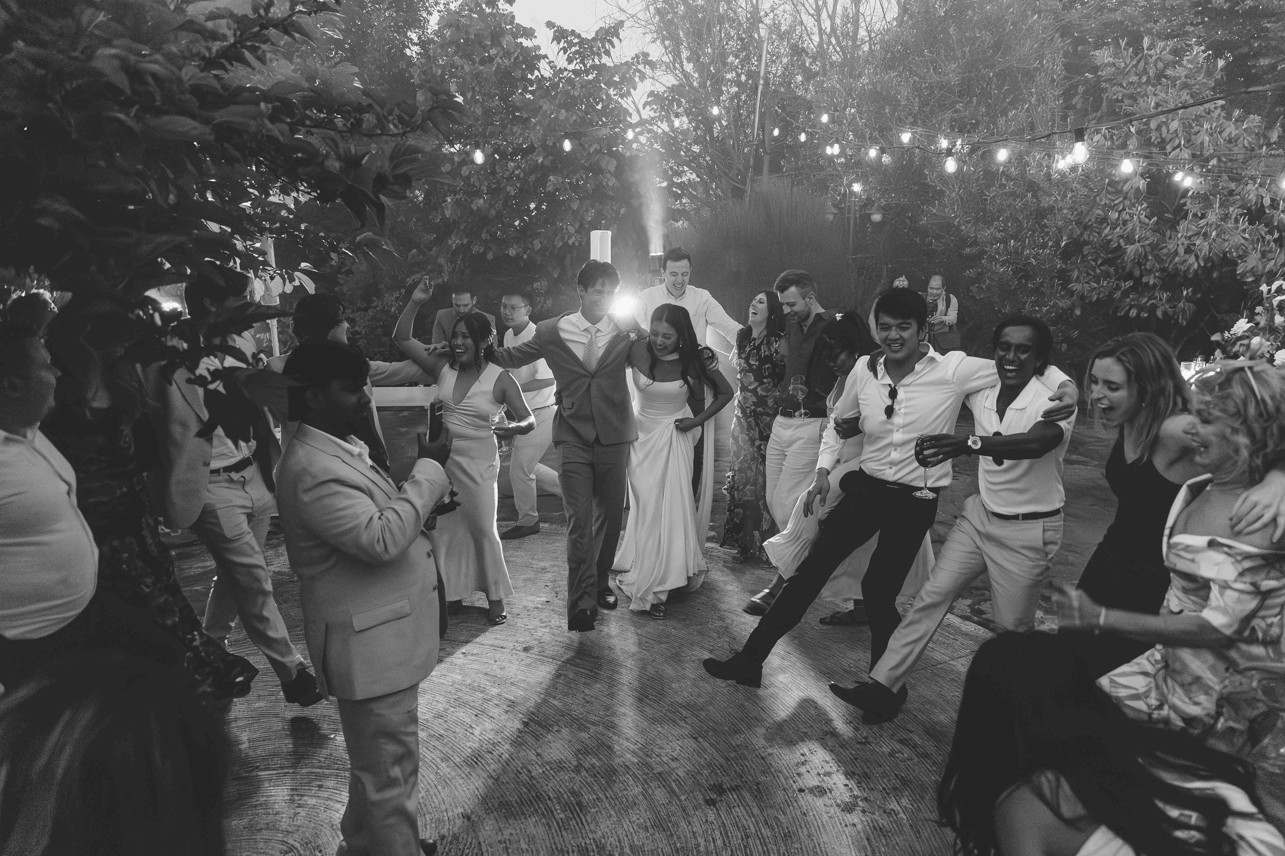 People dancing and celebrating outdoors at a party with string lights overhead, some holding drinks, some capturing photos, and a sprinkler in the background