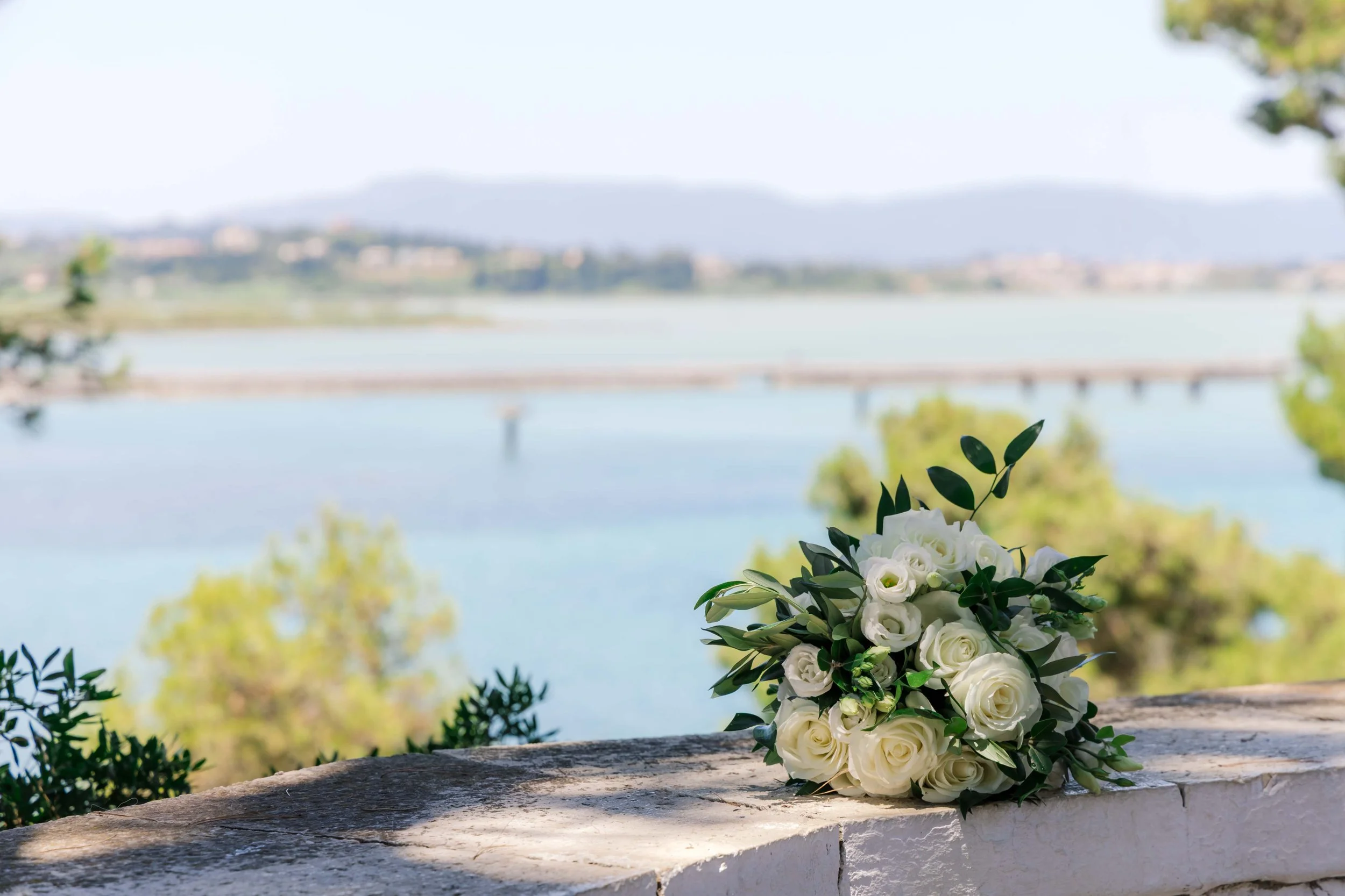 A bouquet of white roses and greenery placed on a stone ledge with a scenic view of a lake, trees, and distant hills in the background.
