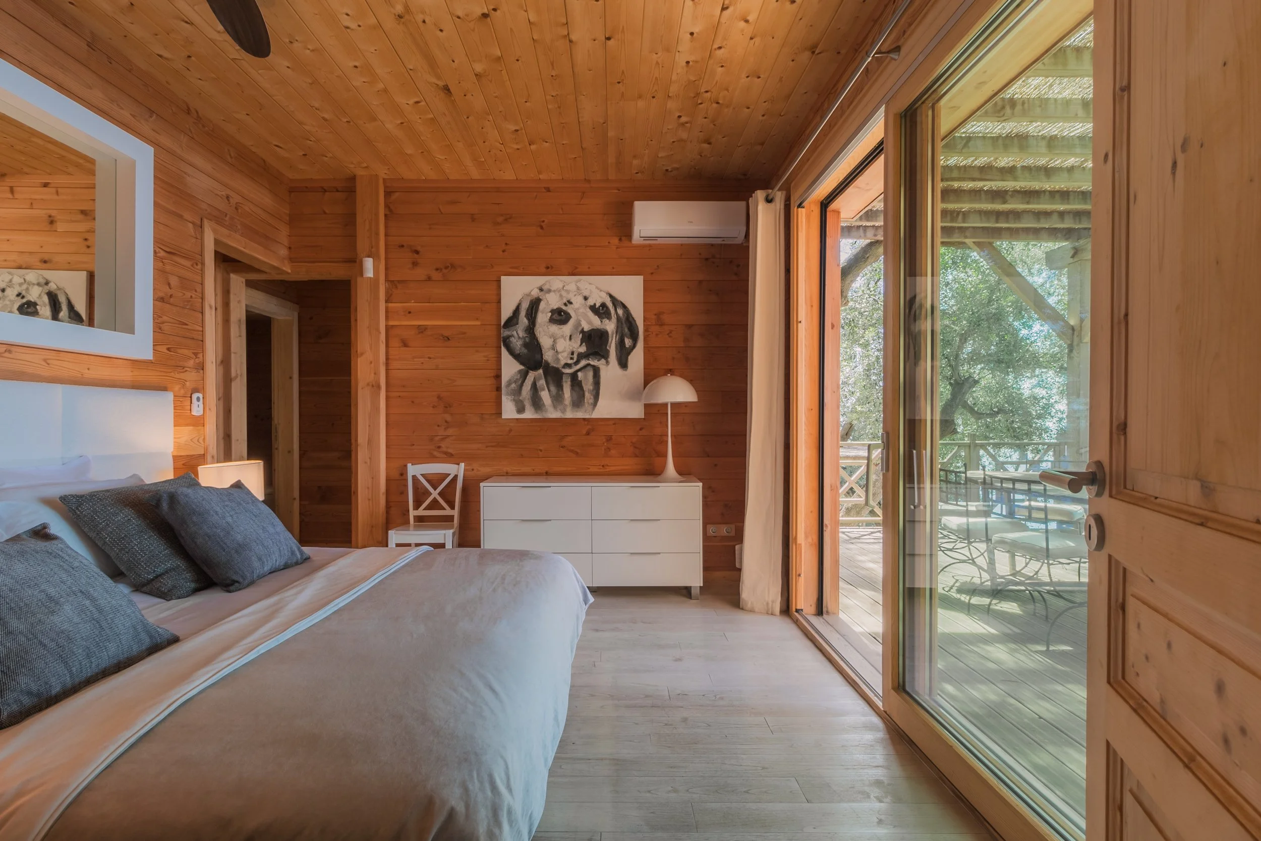 Bedroom with wooden walls, a bed with gray pillows, a white dresser with a lamp, a painting of a dog on the wall, and sliding glass doors leading to an outdoor patio.