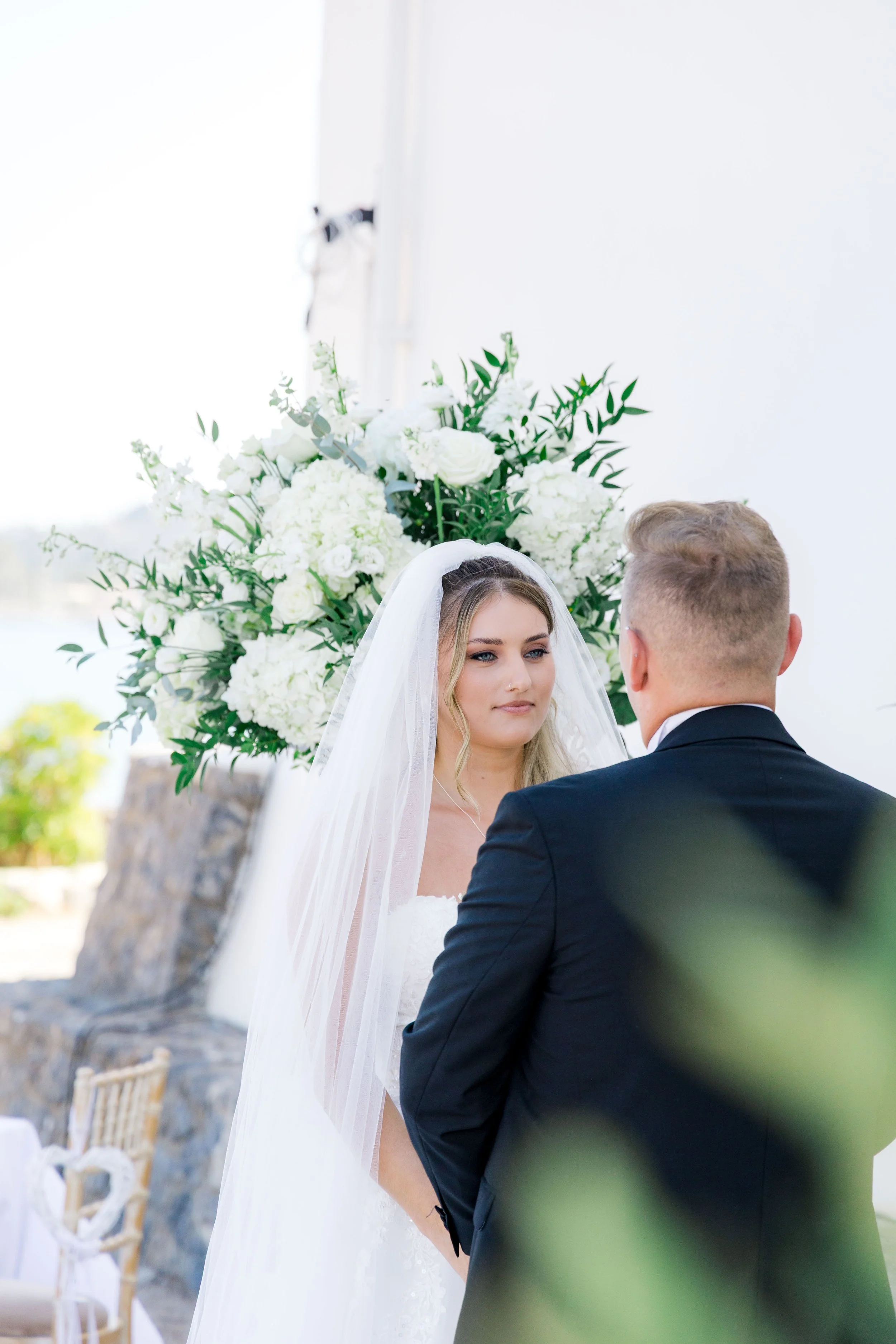 Bride and groom during their wedding ceremony, with a large floral arrangement of white flowers and greenery in the background.