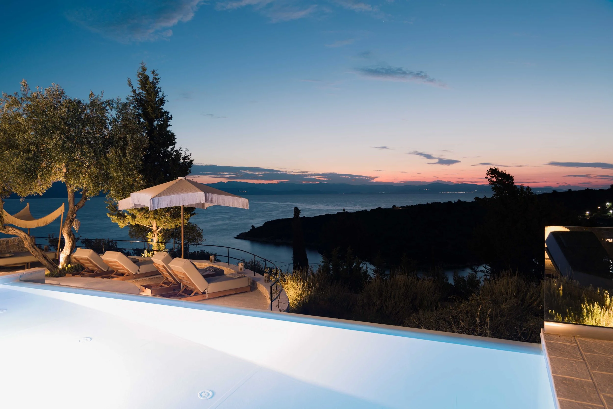 A luxurious outdoor pool area overlooking a body of water, with lounge chairs, umbrellas, and trees at sunset.