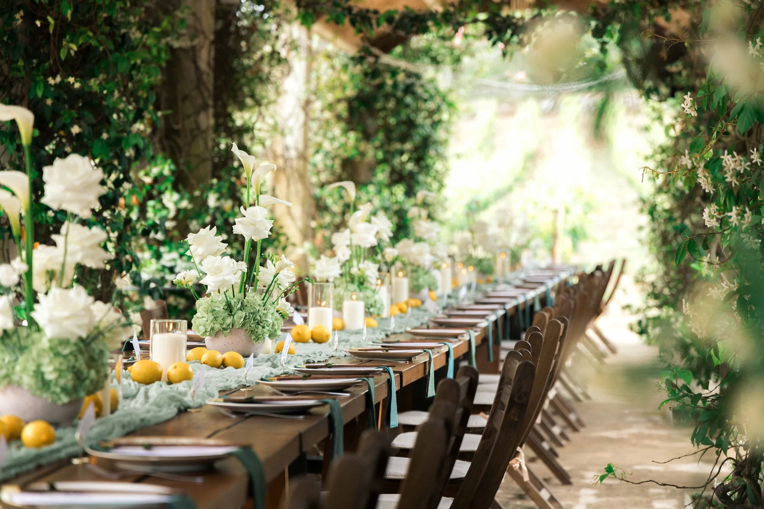 Long outdoor dining table decorated with white flowers, candles, and lemons in a lush, green garden setting.
