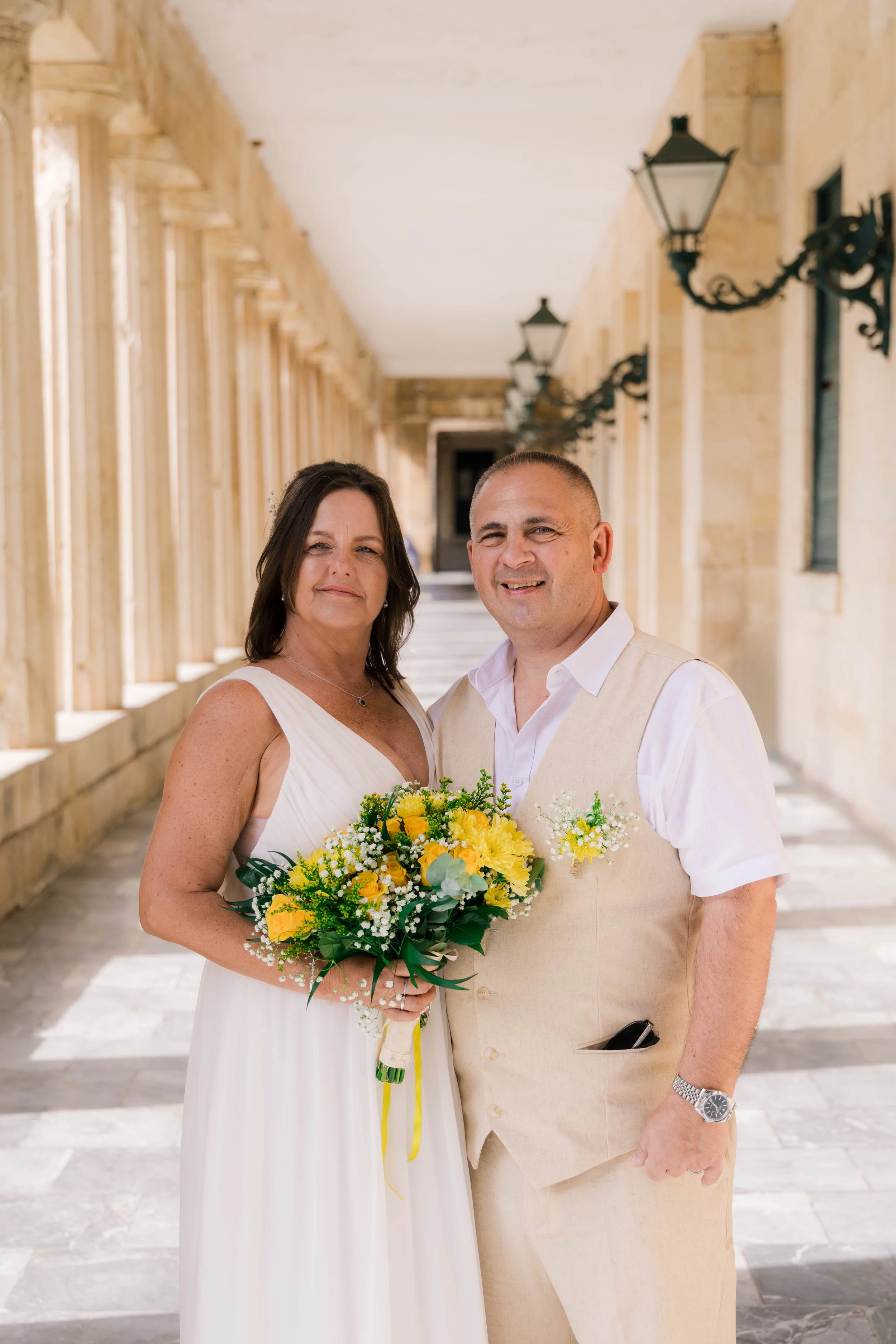 A bride and groom standing together in an outdoor corridor, smiling, with the bride holding a bouquet of yellow and white flowers.
