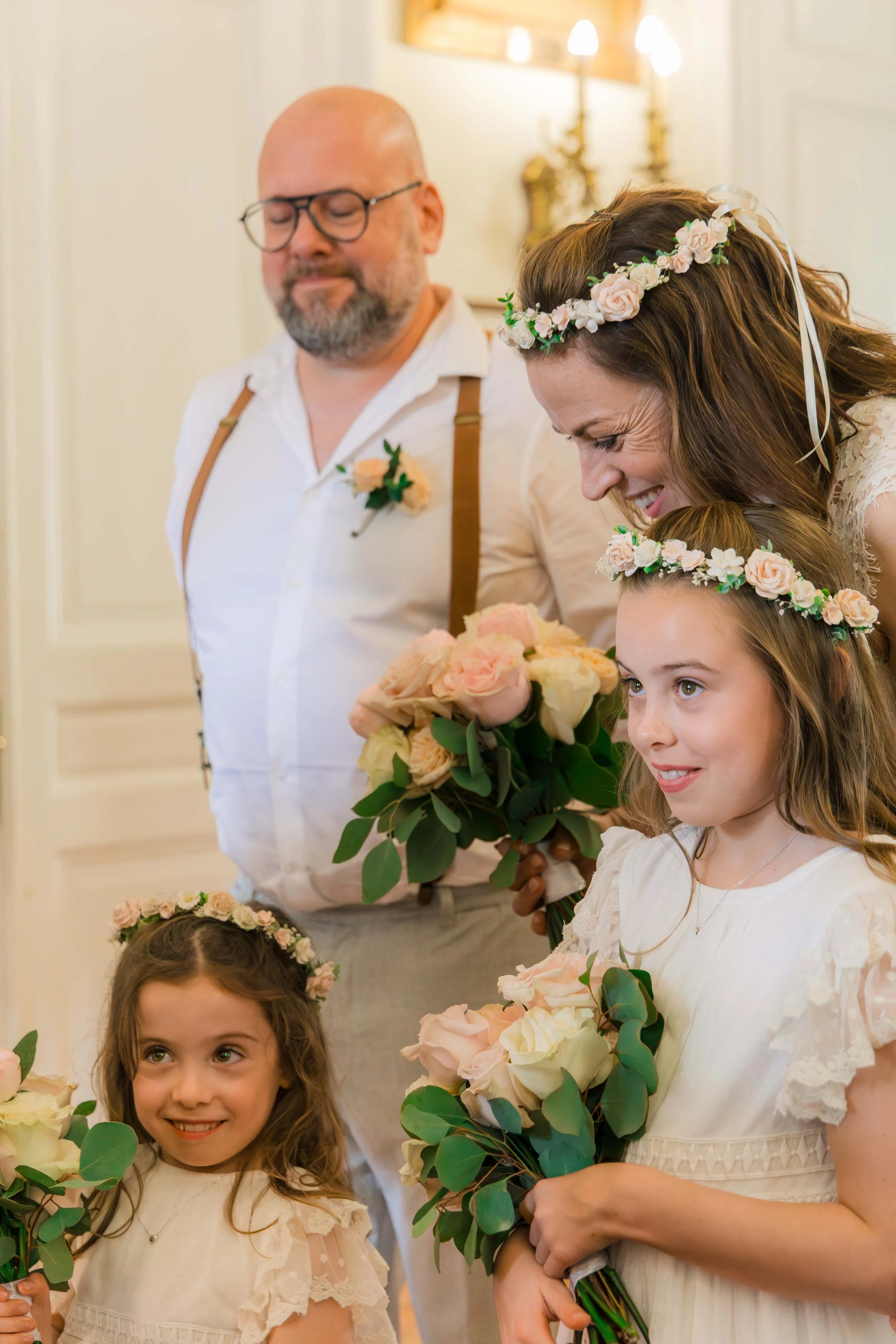 A bride, groom, and two young girls at a wedding, all wearing floral crowns and holding bouquets of roses and greenery, inside a decorated room.