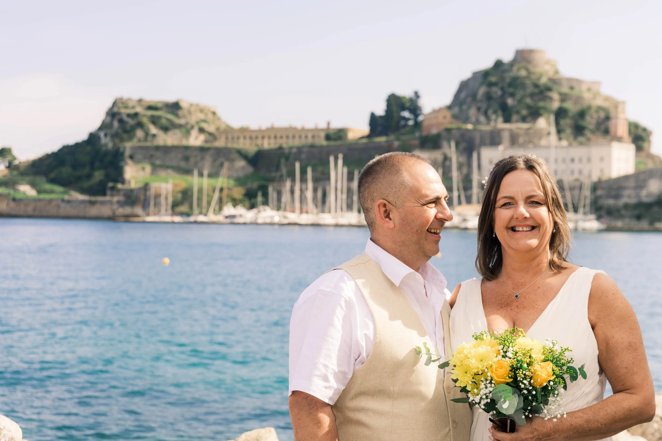 A smiling couple standing near the water, with a castle on a hill and sailboats in the background.