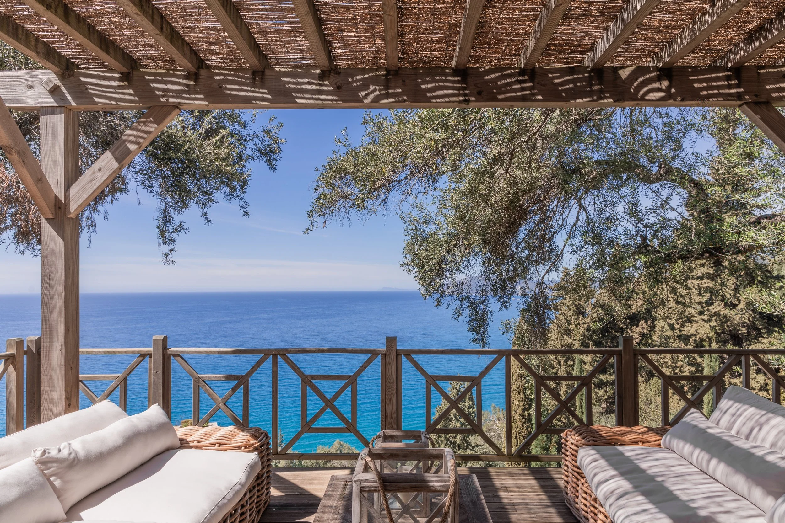 A wooden deck with outdoor furniture, overlooking the ocean, shaded by trees and a bamboo roof.