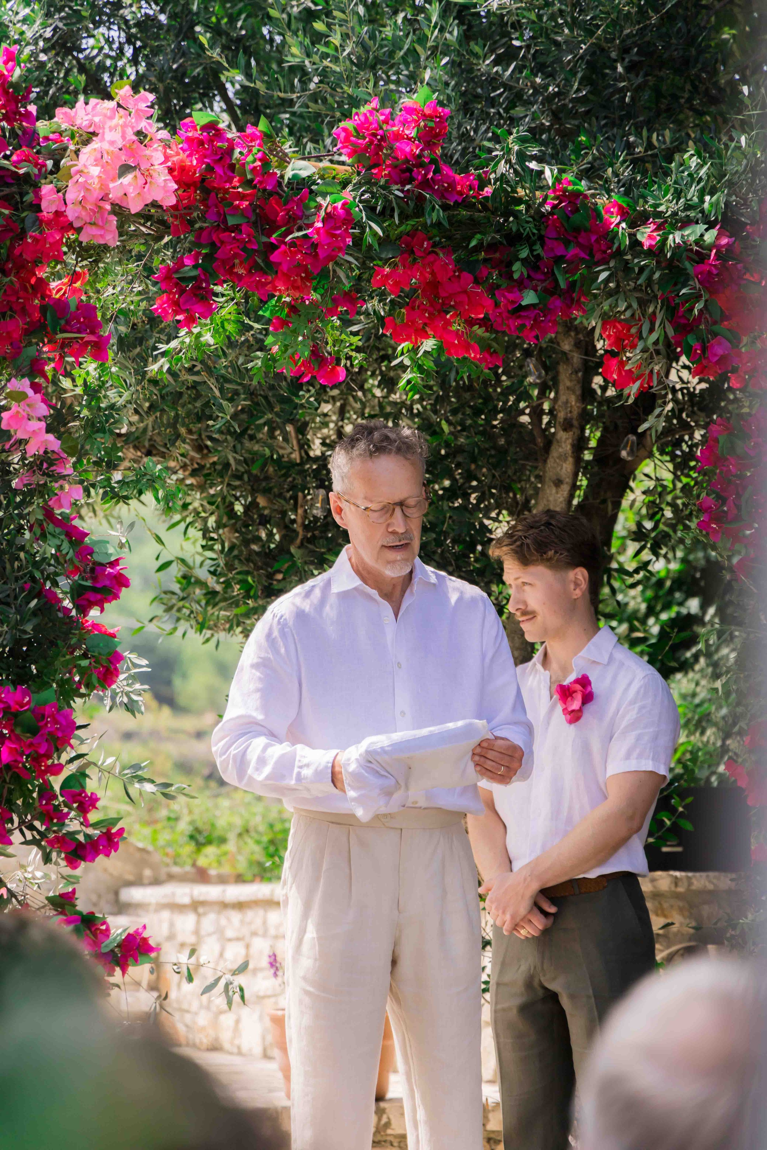 An outdoor wedding ceremony with a man and a young man standing under a flowering bougainvillea arch. The man is reading from a paper and wearing a white shirt with a white glove, while the young man is dressed in a white shirt with a pink flower pin