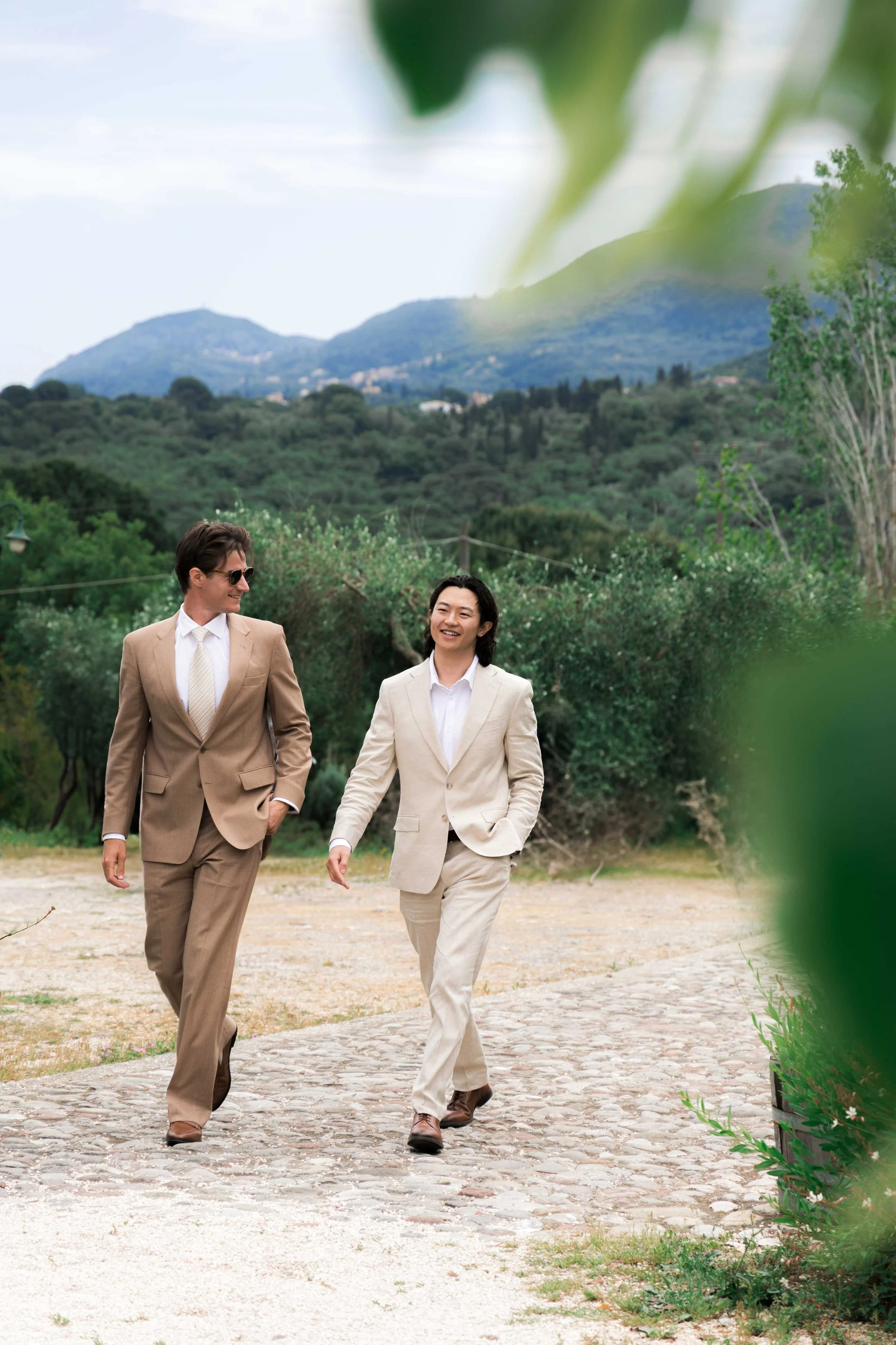 Two people walking outdoors on a cobblestone path, dressed in beige suits, smiling and conversing, with green trees, hills, and mountains in the background.