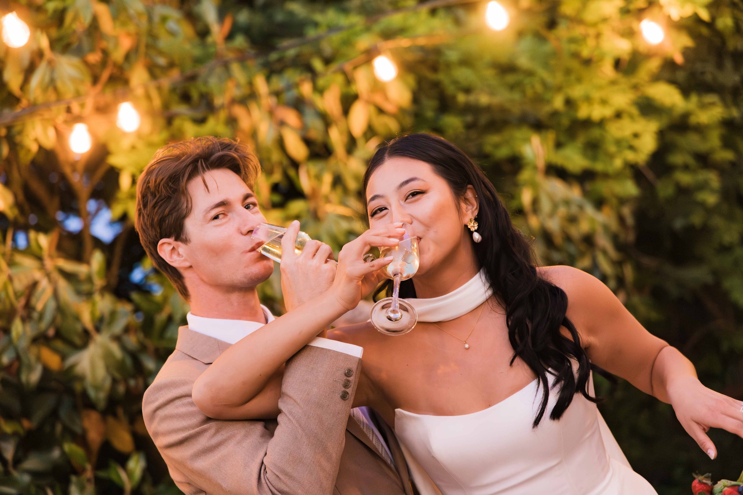 A young man in a beige suit and a young woman in a white dress are enjoying glasses of white wine together at an outdoor event illuminated by string lights and surrounded by greenery.