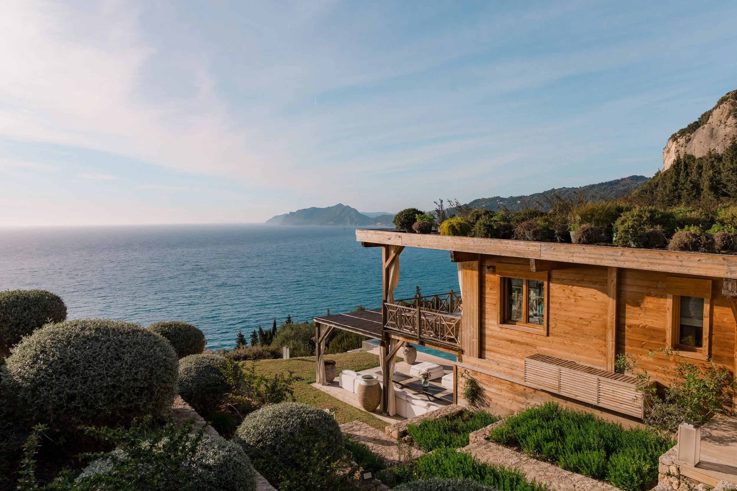 A wooden house with a patio overlooking the ocean, lush greenery, and distant mountains under a partly cloudy sky.