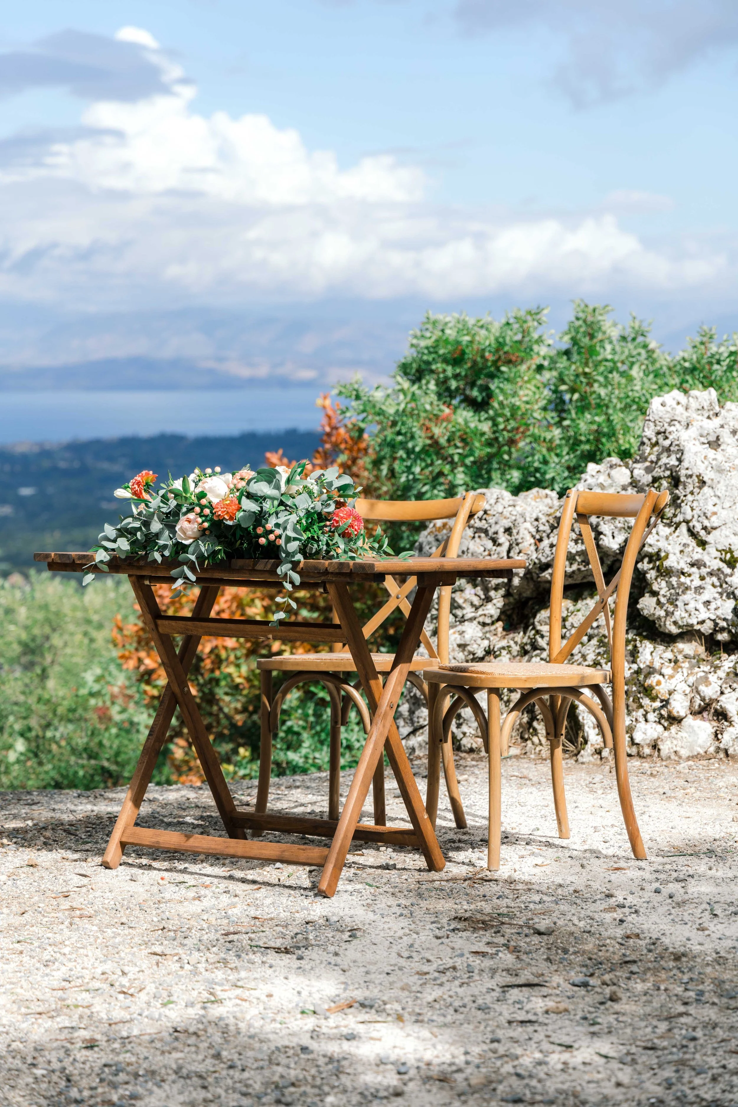 A wooden table with a floral centerpiece and two wooden chairs outdoors on a gravel surface, with green bushes, rocks, and a distant body of water under a partly cloudy sky.