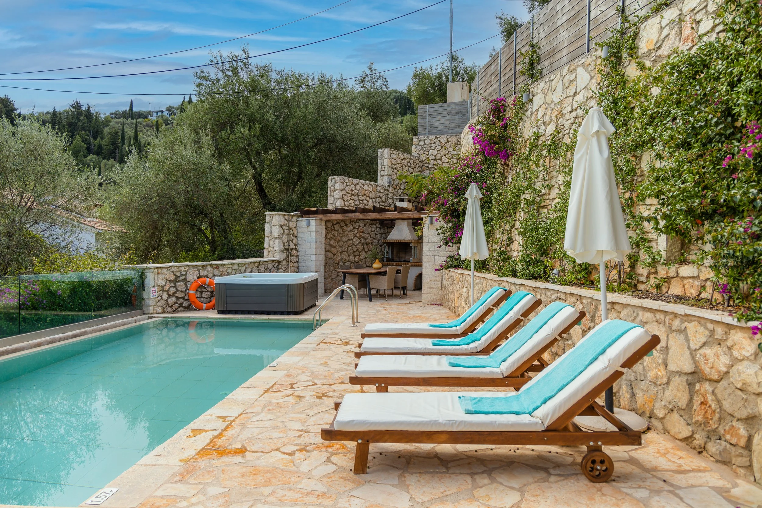 Poolside scene with four white lounge chairs and blue towels, two white umbrellas, a hot tub, and an outdoor kitchen with a fireplace, surrounded by stone walls and greenery, under a partly cloudy sky.