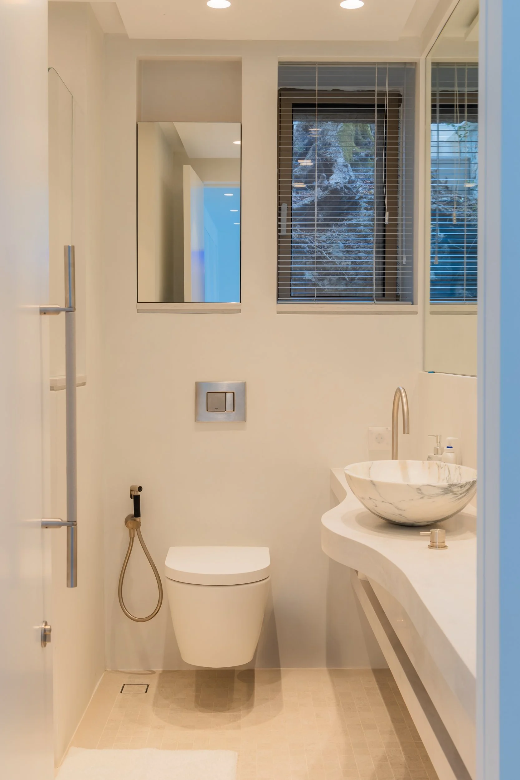 Minimalist bathroom with a wall-mounted toilet, a marble sink basin, and a large mirror, with windows showing outdoor scenery.