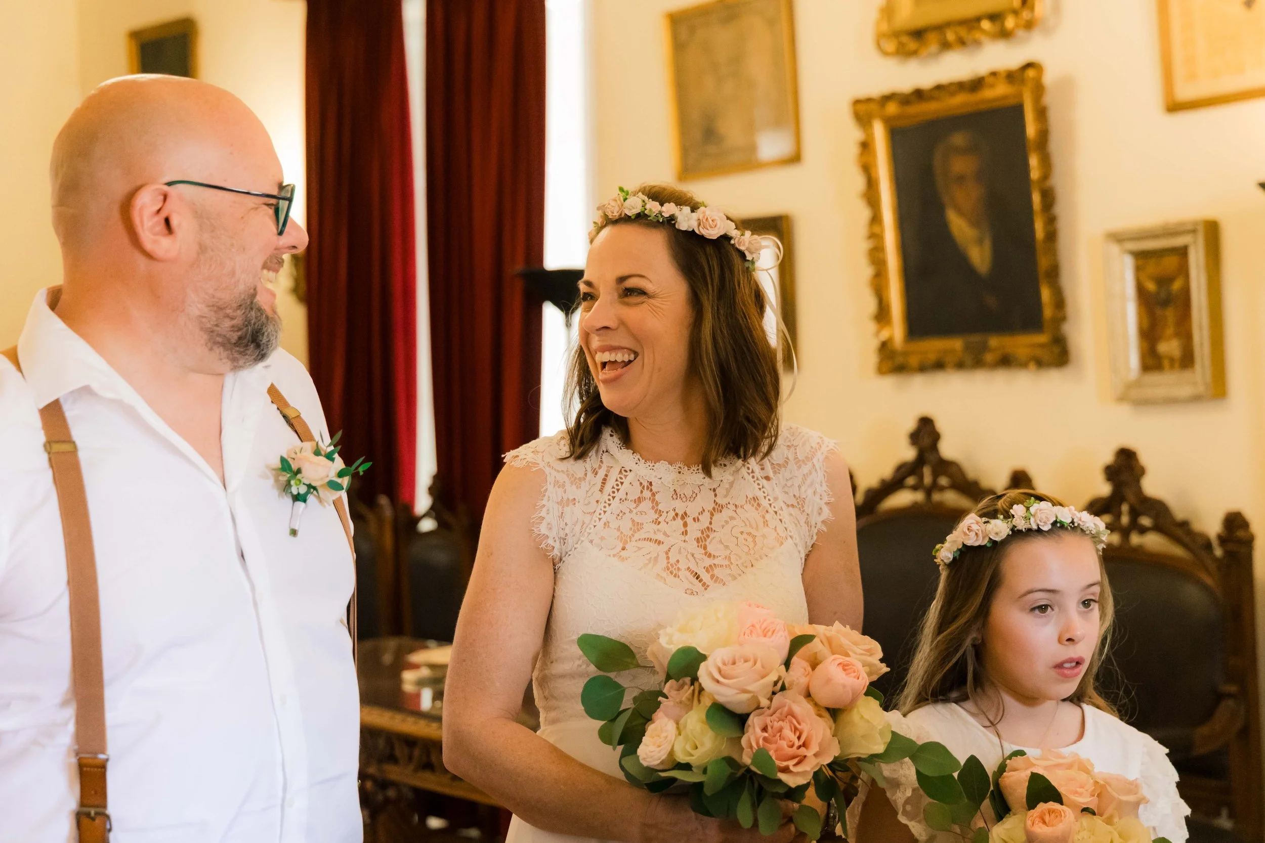A woman in a white lace dress holding a bouquet of pink and white roses, smiling at a man in glasses and a white shirt, with a young girl in a white dress and floral crown also holding a bouquet, in a warmly decorated room.