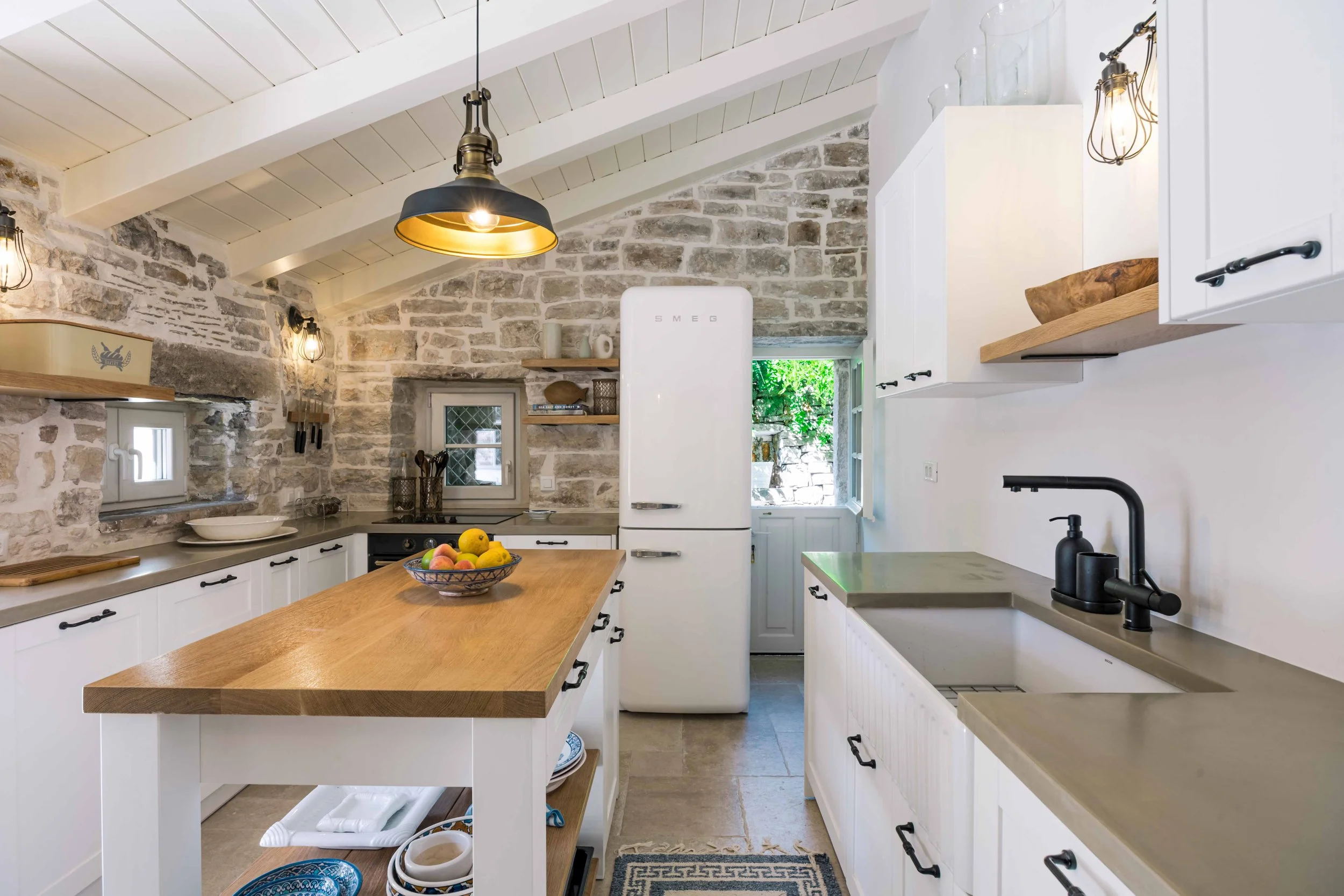 A rustic kitchen with white cabinets, a wooden island with fruit, a SMEG refrigerator, stone walls, black pendant lights, and open shelves with glass jars and bowls.
