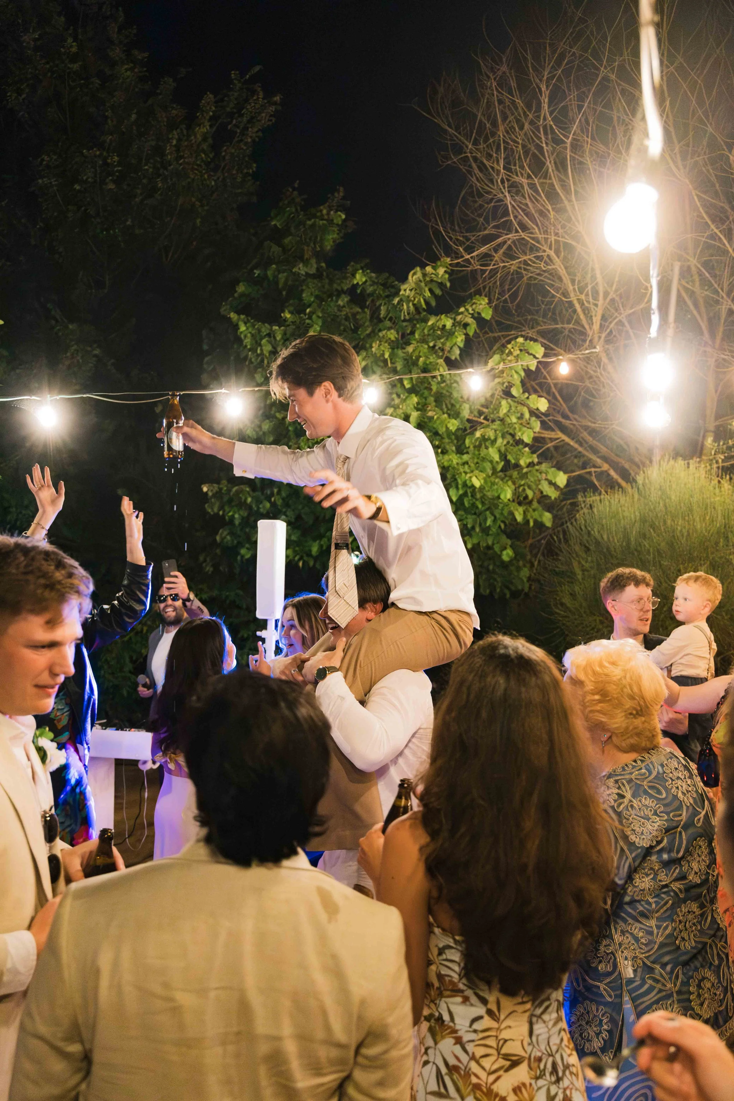 People celebrating at an outdoor party at night with string lights overhead. A man is on someone’s shoulders holding a beer and smiling, surrounded by others talking, dancing, and taking photos.