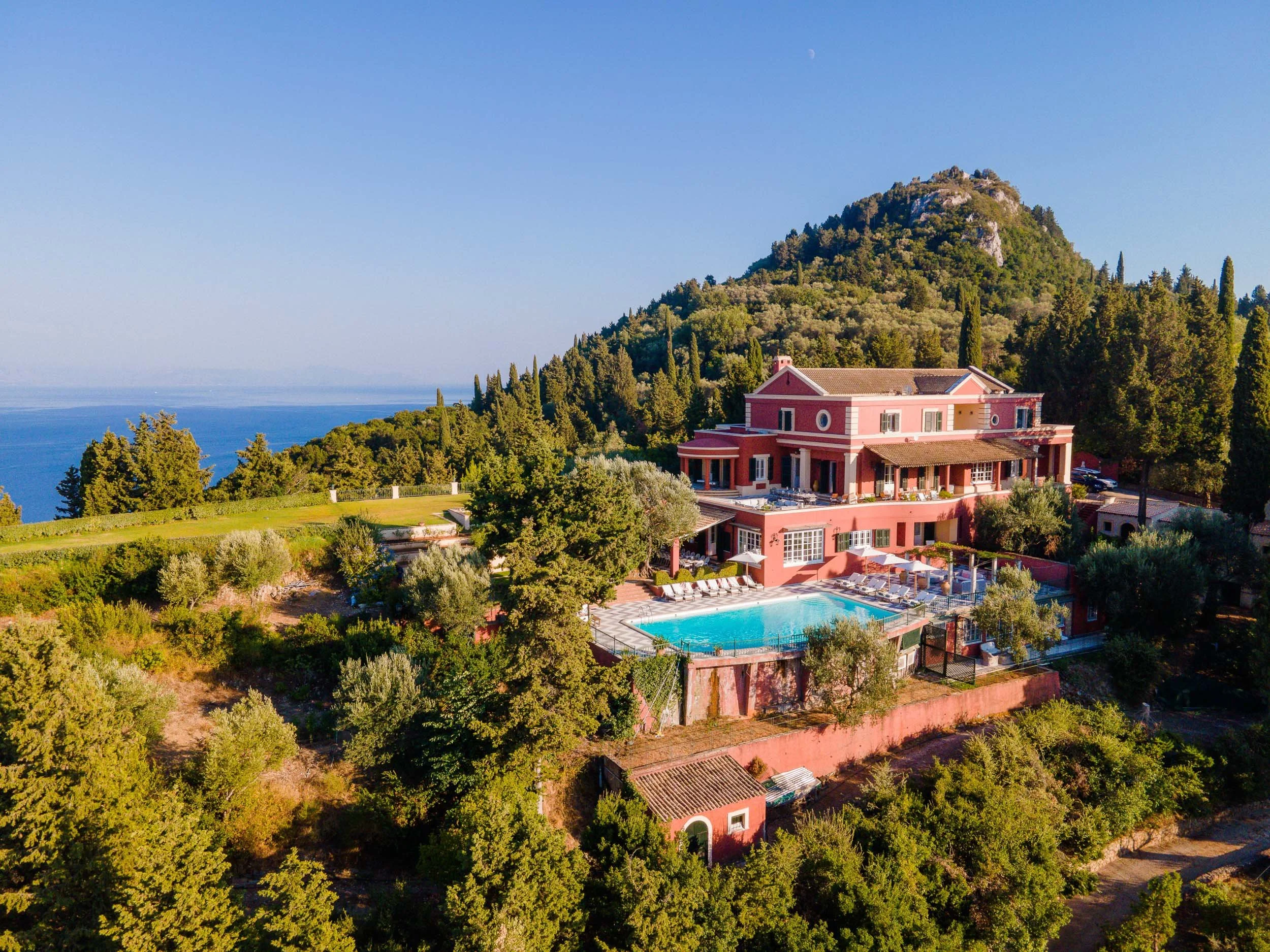 A pink multi-story house with a swimming pool surrounded by trees and lush greenery, set on a hillside with mountains in the background and the sea in the distance.