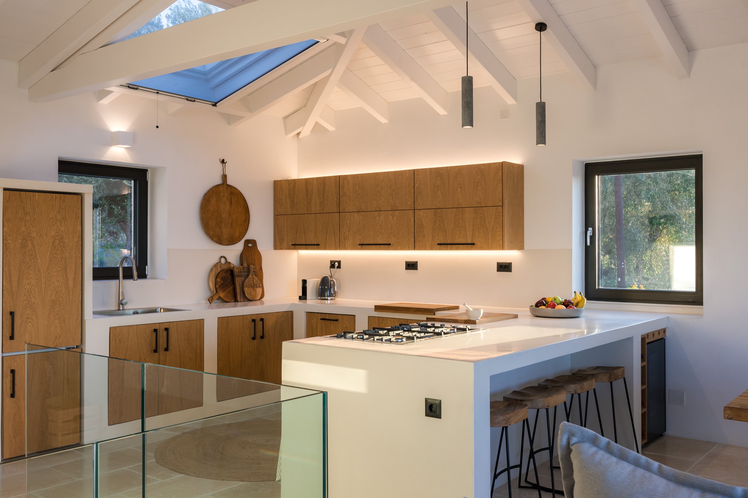Modern kitchen with wooden cabinets, white countertops, and large windows. Skylight above, pendant lights hanging from white vaulted ceiling with exposed beams. Fruit bowl on counter, cutting boards and kettle on wall, and bar stools at breakfast bar