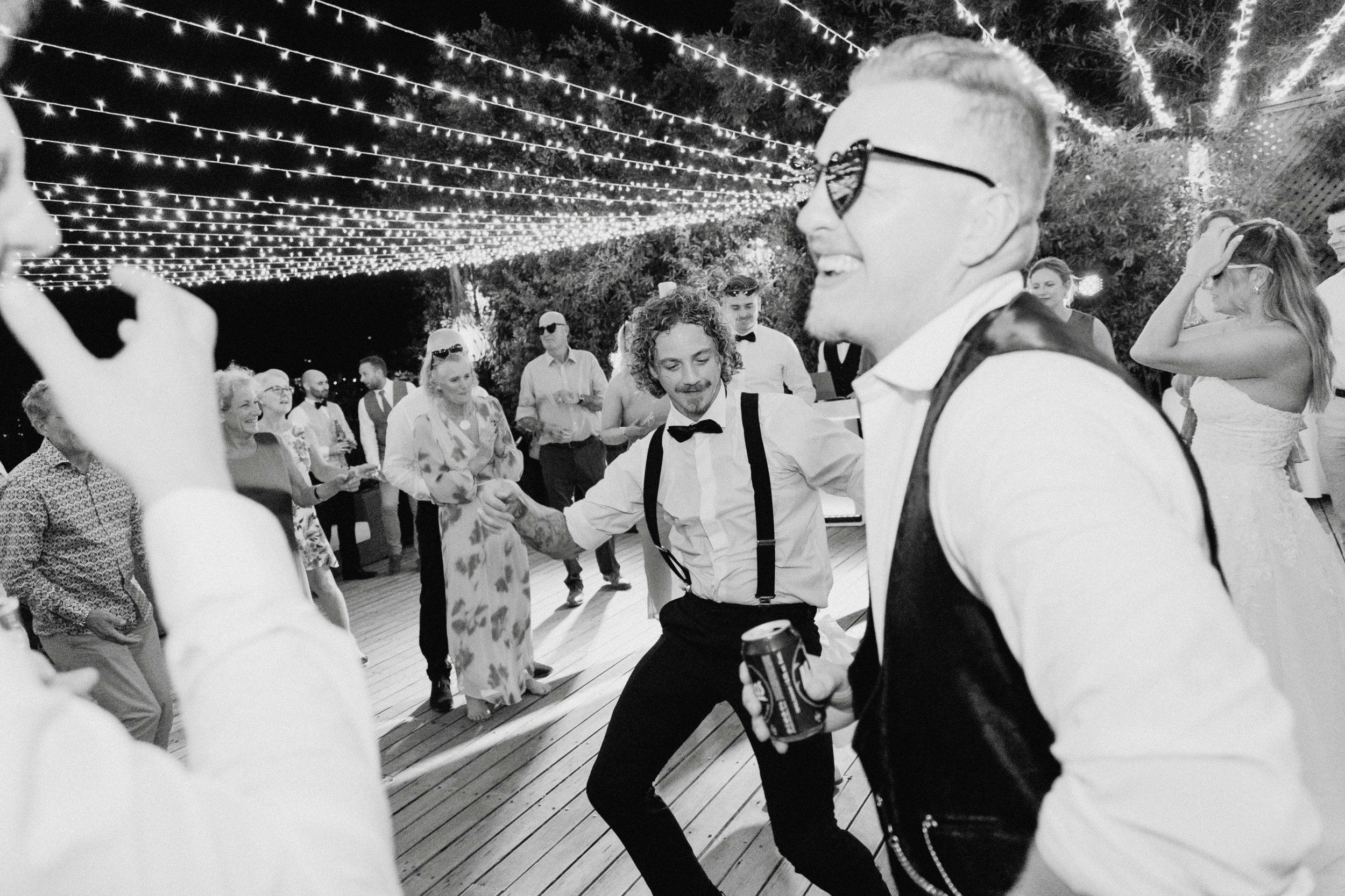 People dancing at a party outdoors at night, decorated with string lights, with a mix of ages and dressed in formal and casual attire.