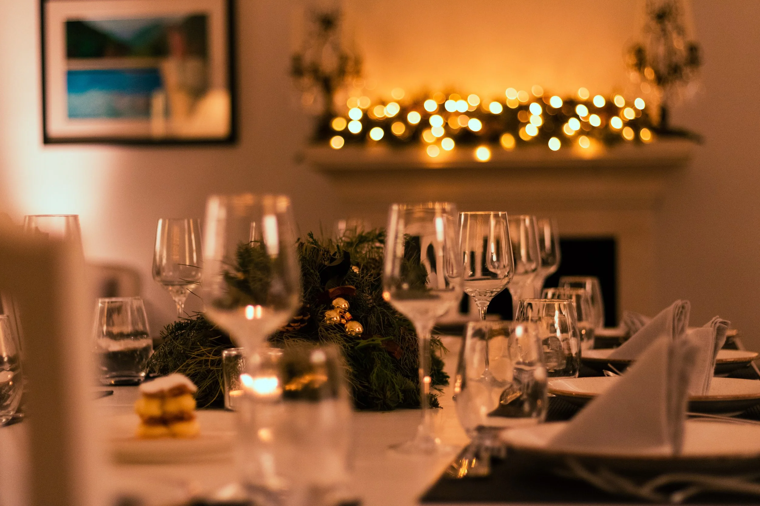 A warmly lit dining table decorated for Christmas with wine glasses, napkins, and a centerpiece with greenery and gold ornaments, with blurred string lights and artwork in the background.