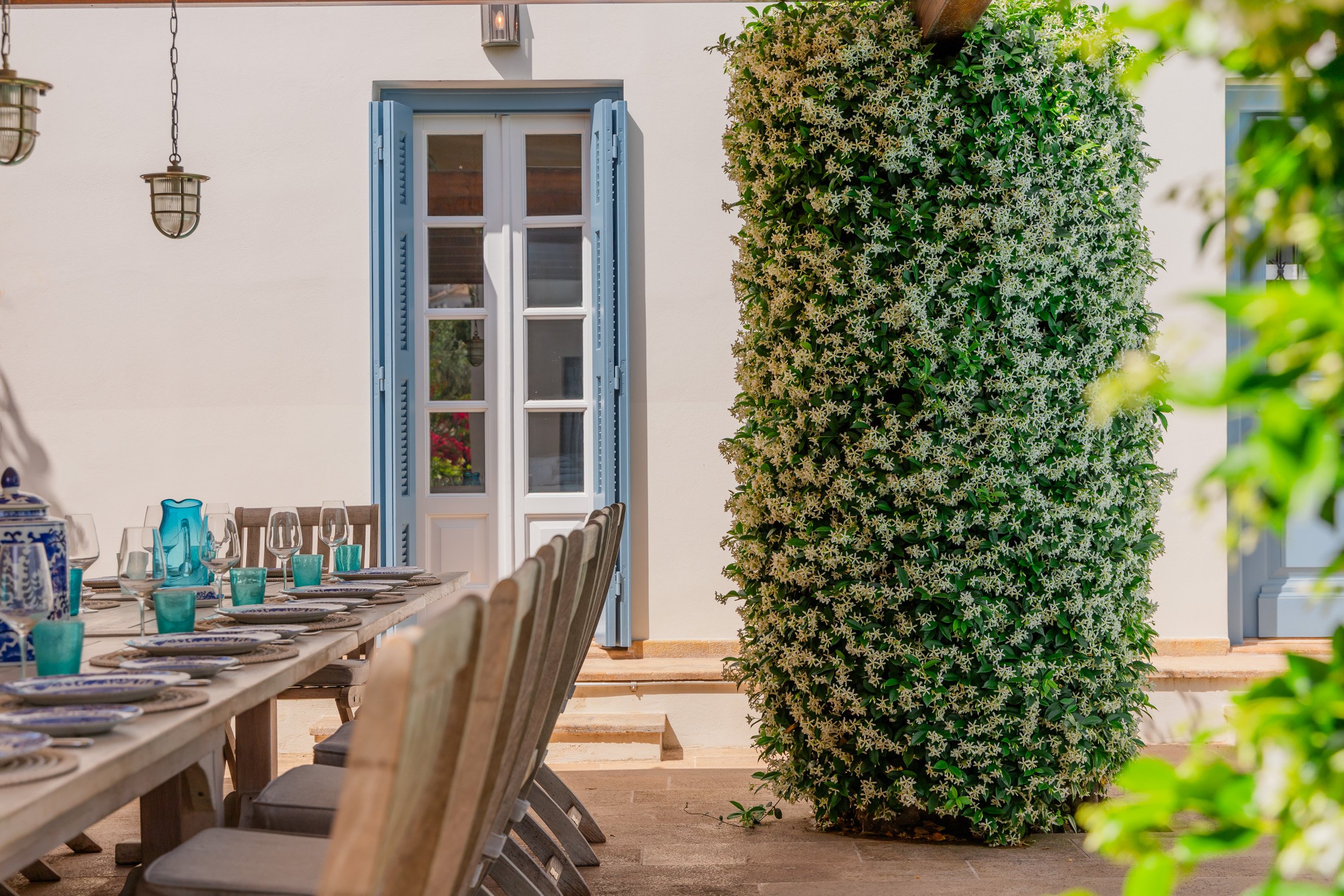 An outdoor dining area with a long wooden table set with glasses, turquoise cups, and plates, positioned in front of a white wall with blue window shutters and a large bush with white flowers on the right side.