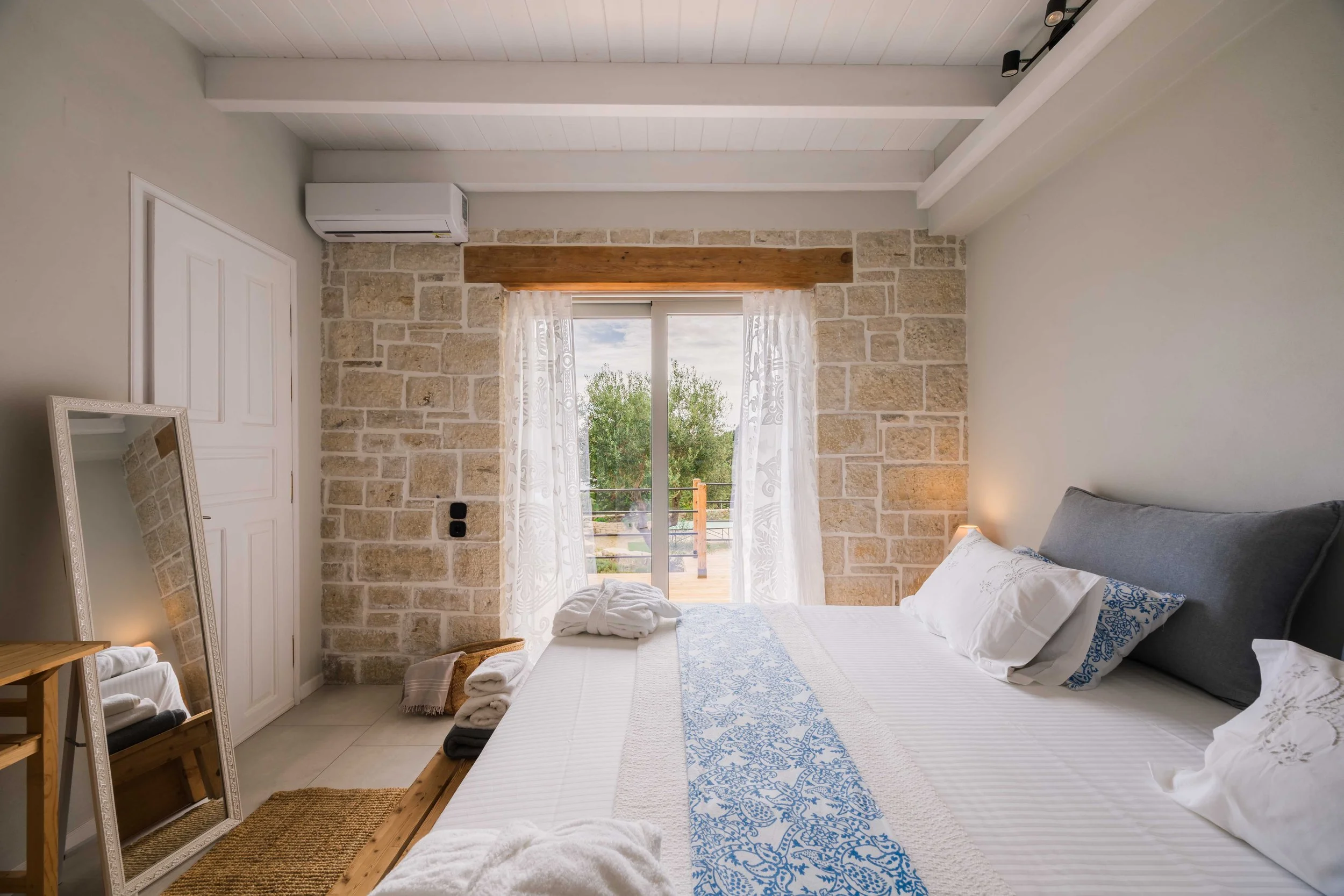 A bedroom with a bed, pillows, and bedding, adjacent to a stone wall with a sliding glass door leading to a balcony with trees outside, and a mirror leaning against the wall.