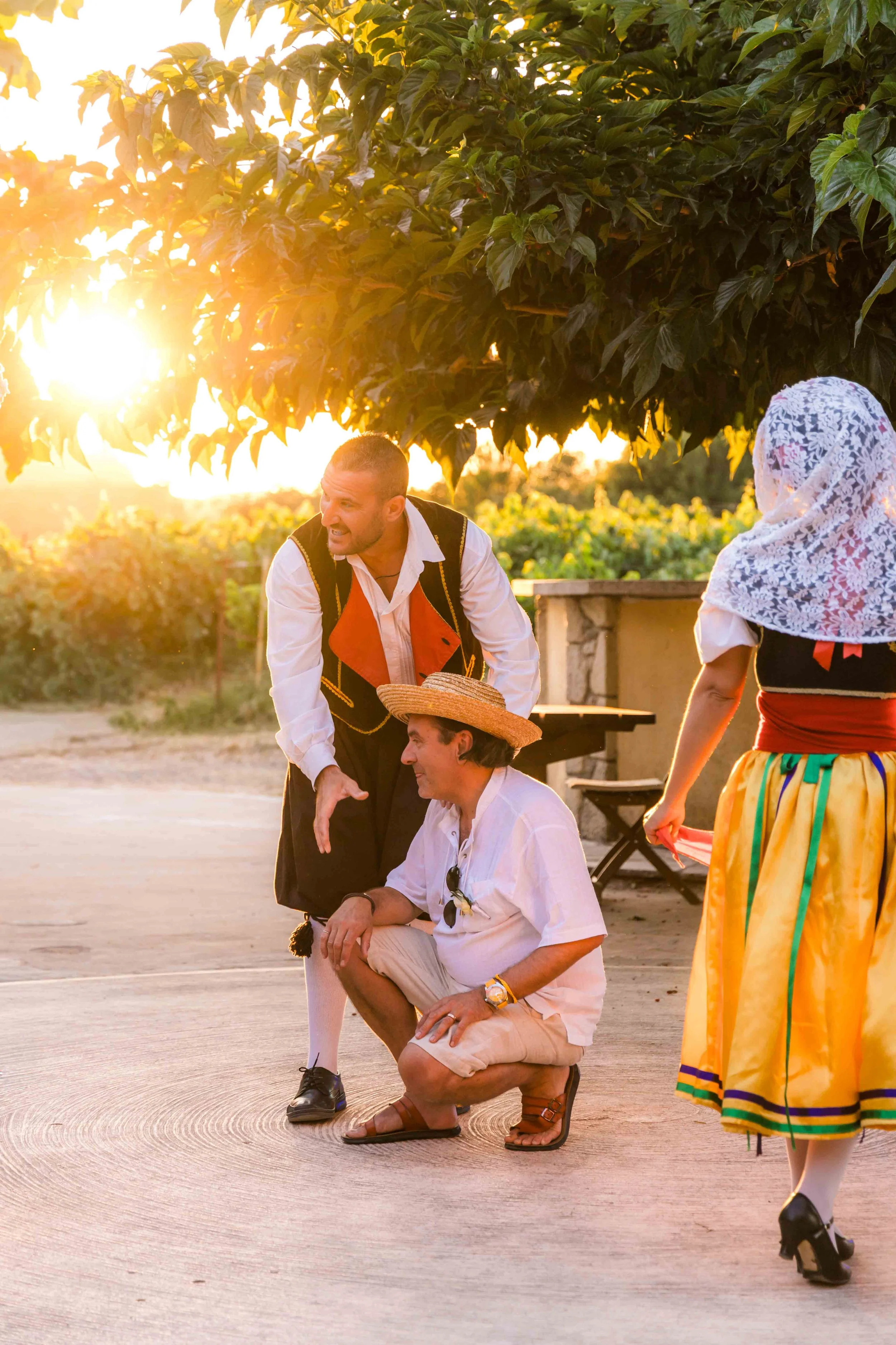 People dressed in traditional Spanish attire, engaging in a conversation outdoors during sunset, with a large tree and vineyard in the background.