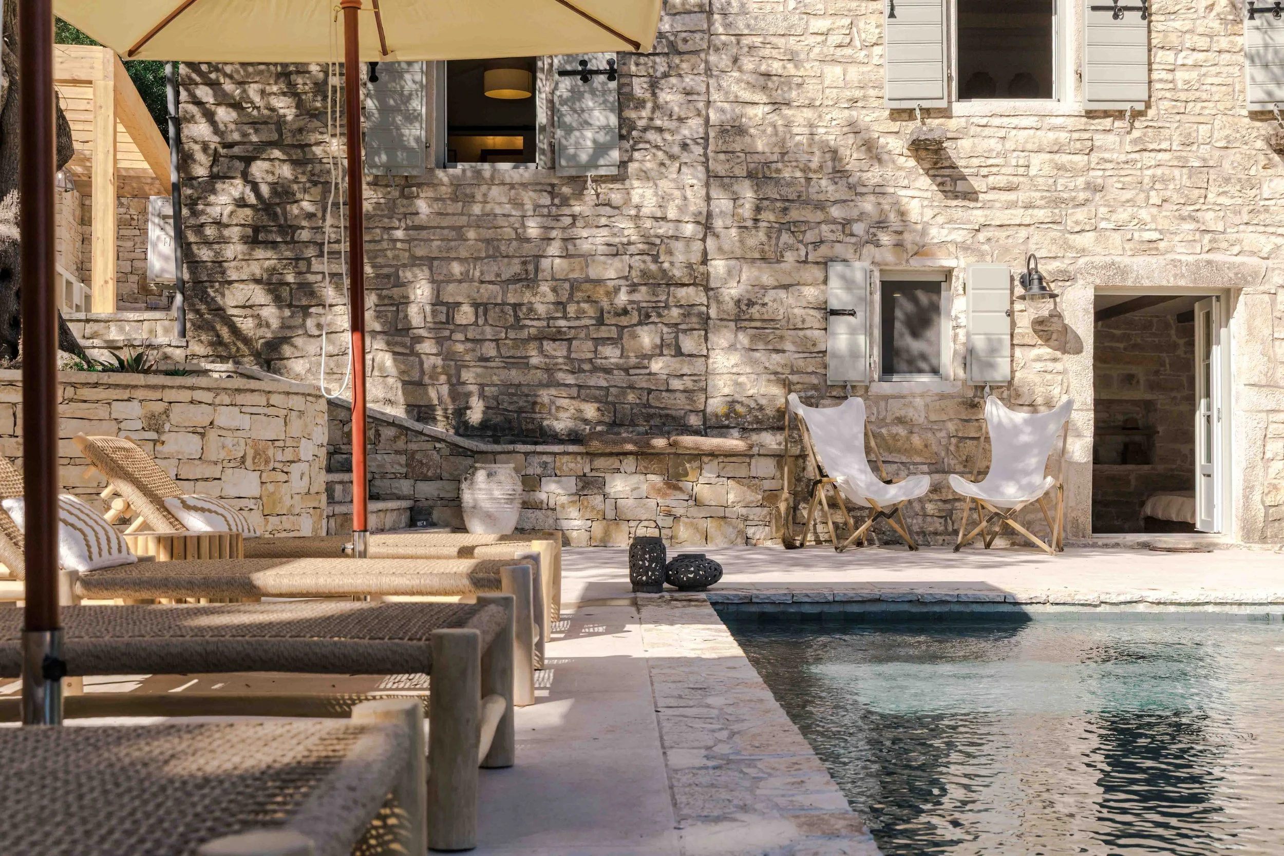 A luxurious outdoor poolside area with sun loungers, a large umbrella, and two white folding chairs next to a stone house with open windows and shutters.