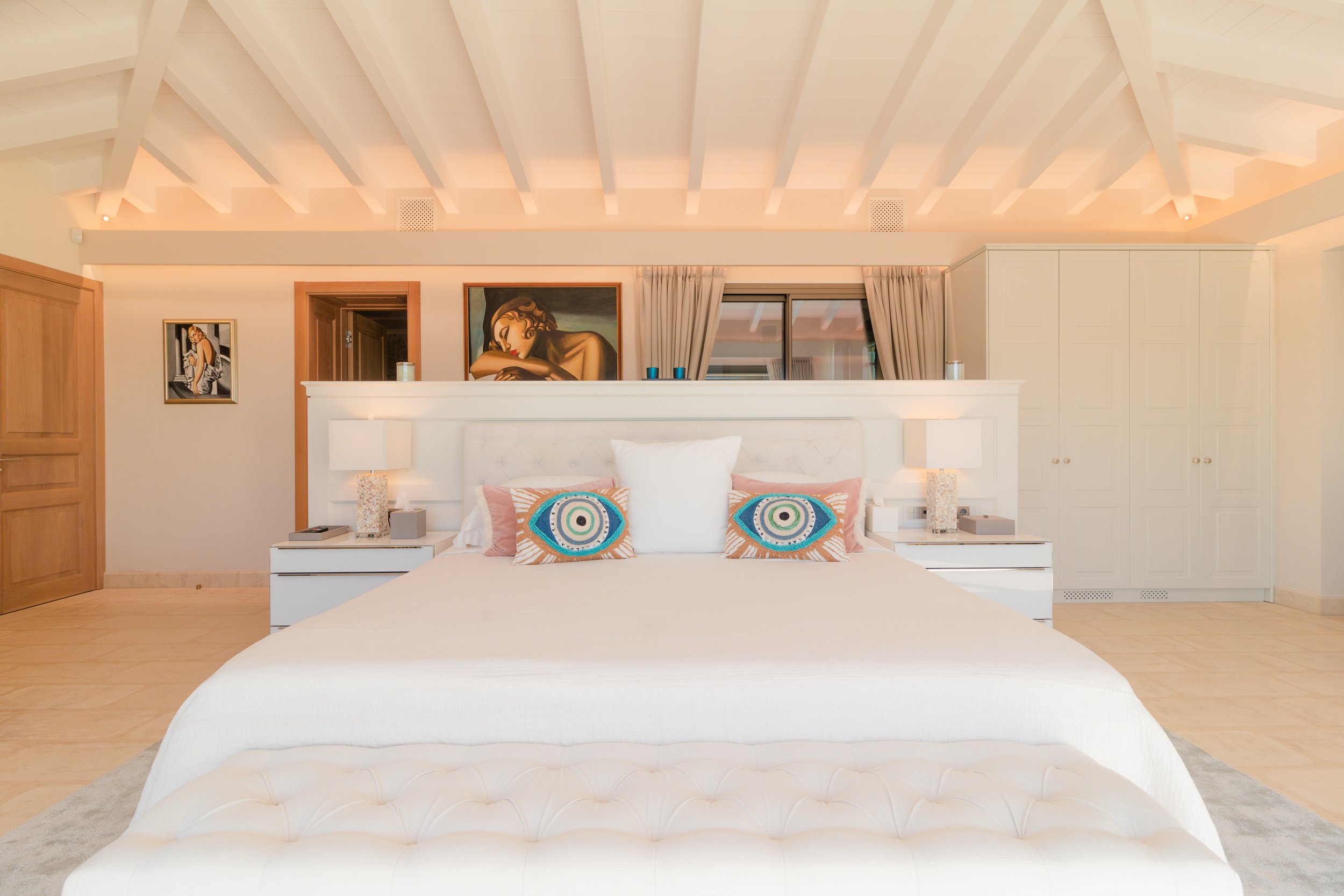 A bedroom with a white bed, colorful pillows, bedside lamps, artwork on the wall, and a storage cabinet.