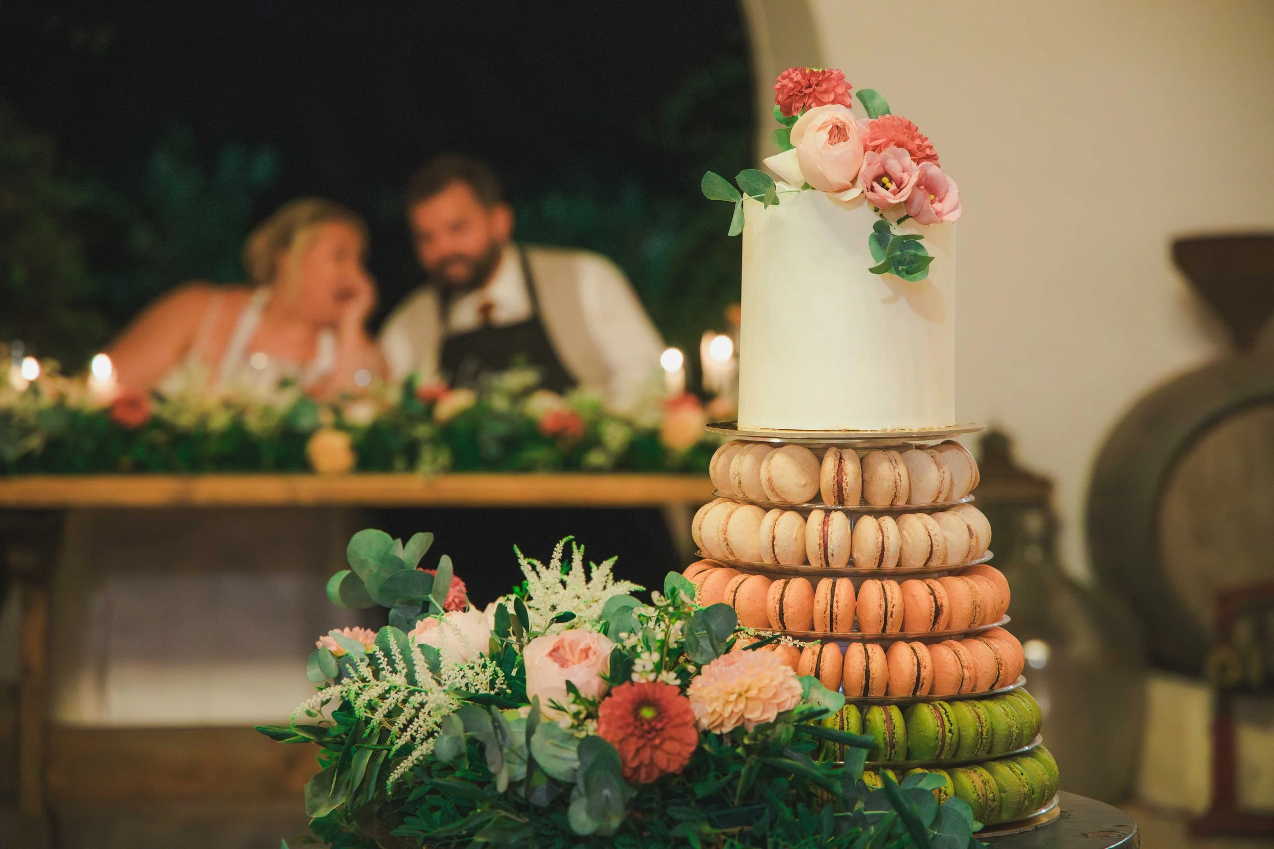 A wedding cake decorated with pink and white flowers on top, surrounded by a bouquet of roses and greenery in the foreground. In the blurred background, a couple is sitting at a table with candles, celebrating at night.