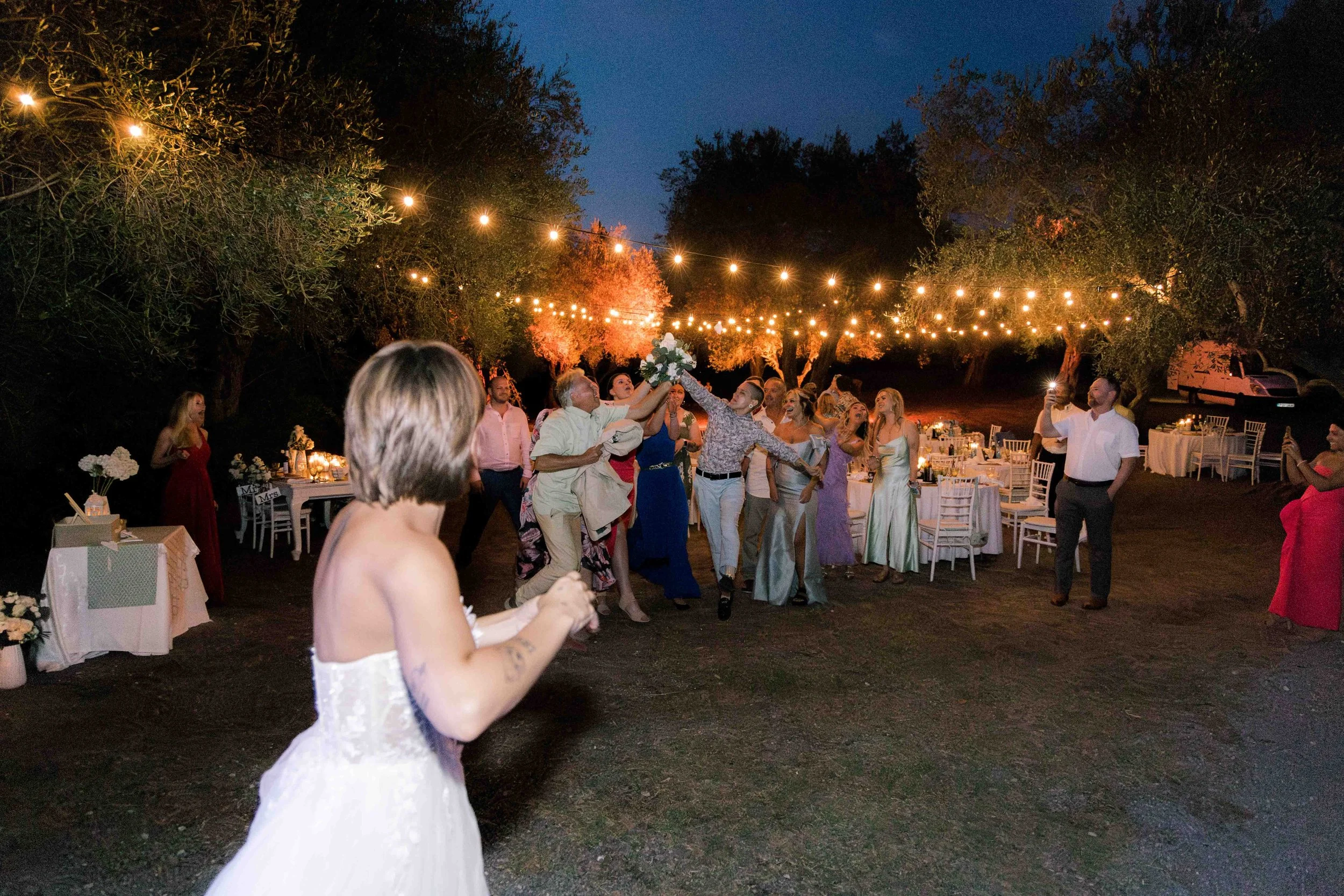 Outdoor wedding reception at night with string lights, guests dancing around bride in white dress, tables decorated with flowers and candles.