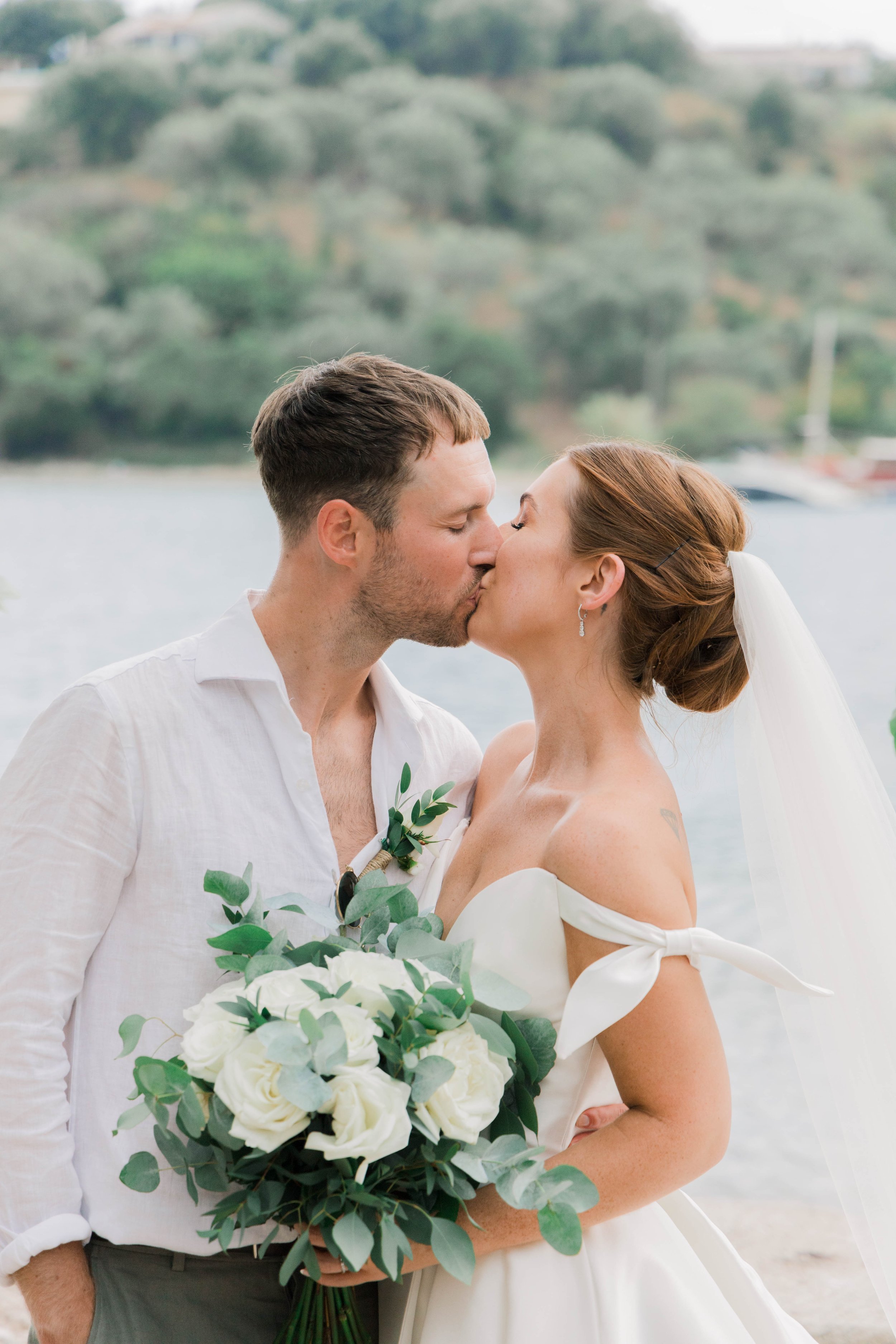 A bride and groom sharing a kiss during their wedding ceremony outdoors by a body of water, with trees and a boat in the background.