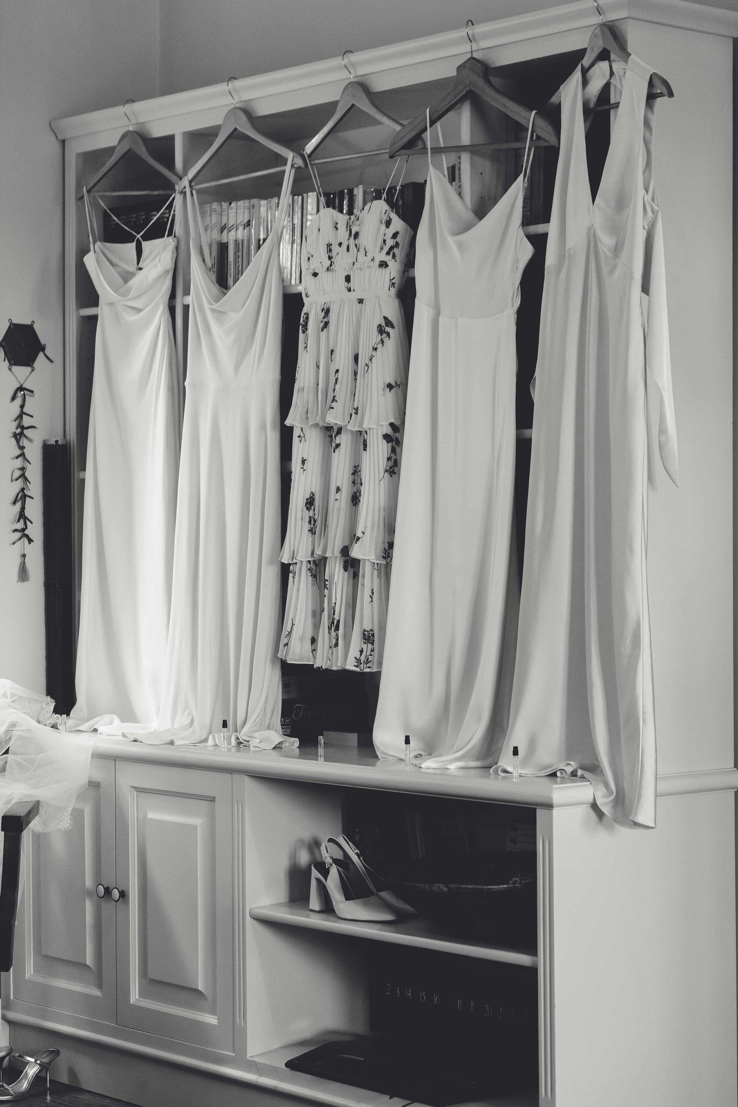 Five white dresses and a patterned dress hanging on hangers in a display case, with a pair of high heels on a lower shelf.