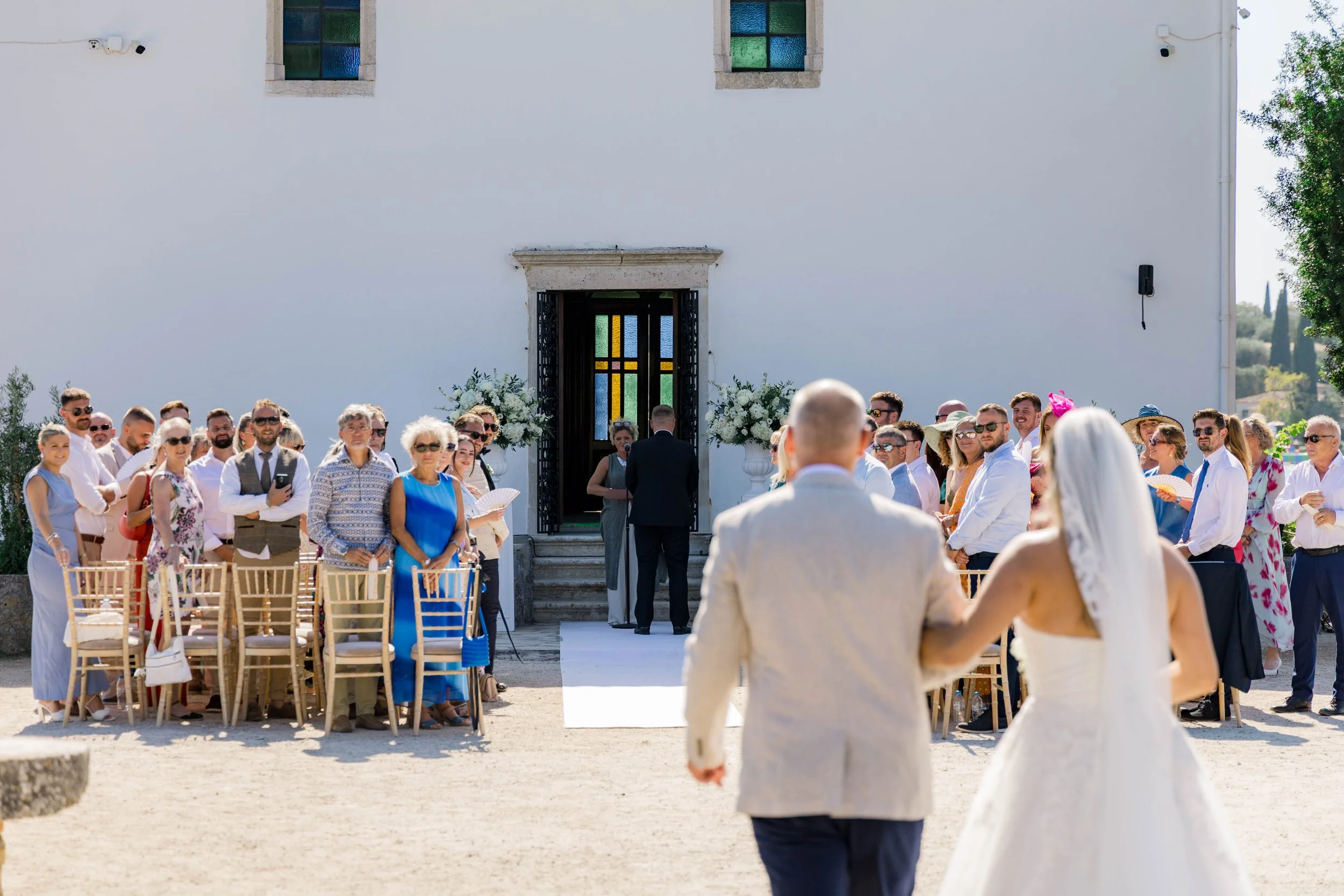 A wedding ceremony taking place outdoors in front of a white building with stained glass windows. The bride and groom are walking hand in hand towards the altar, with guests seated on both sides, some standing and some sitting. The guests are dressed