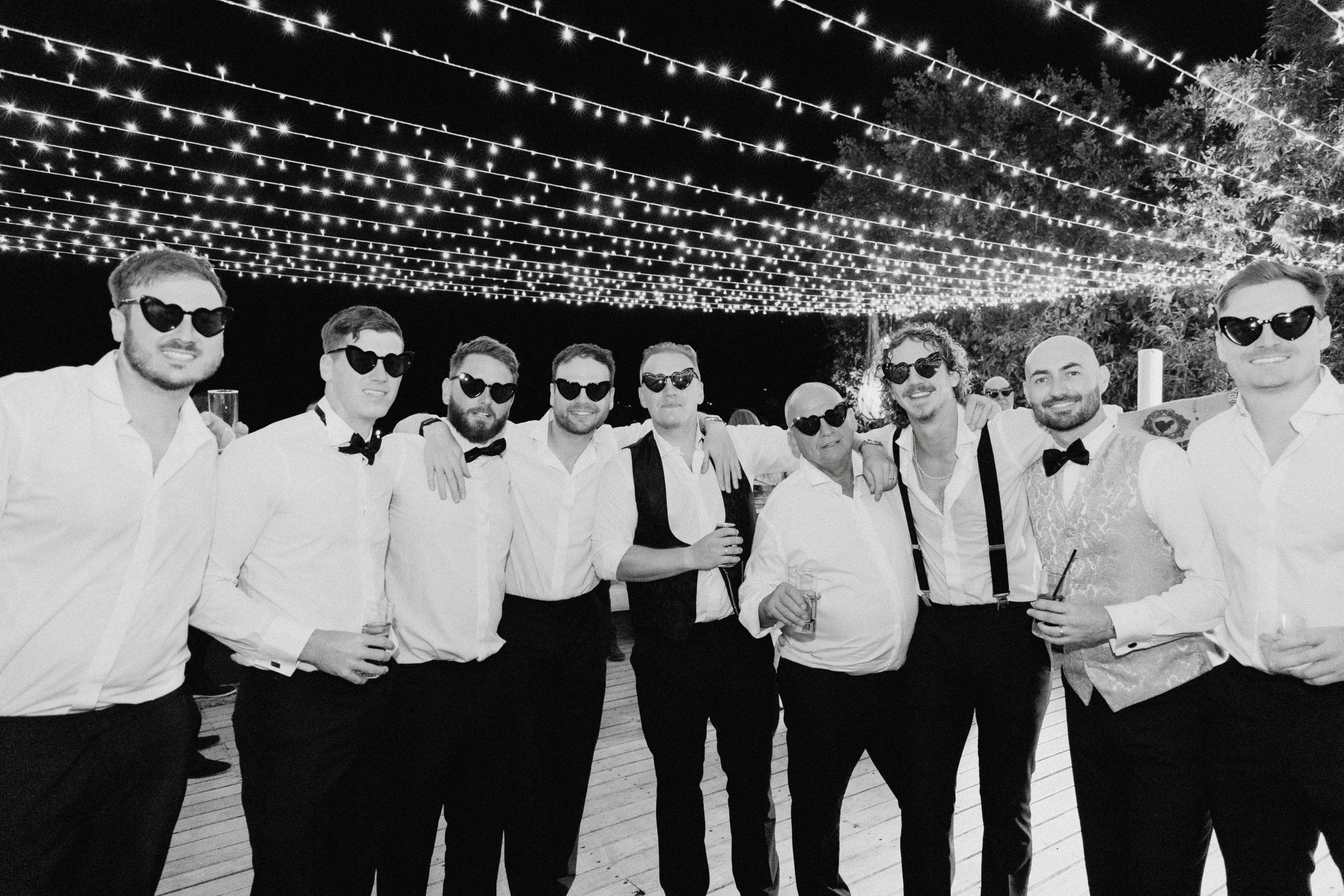 A group of men dressed in formal attire with sunglasses, standing close together at an outdoor evening event with string lights overhead.