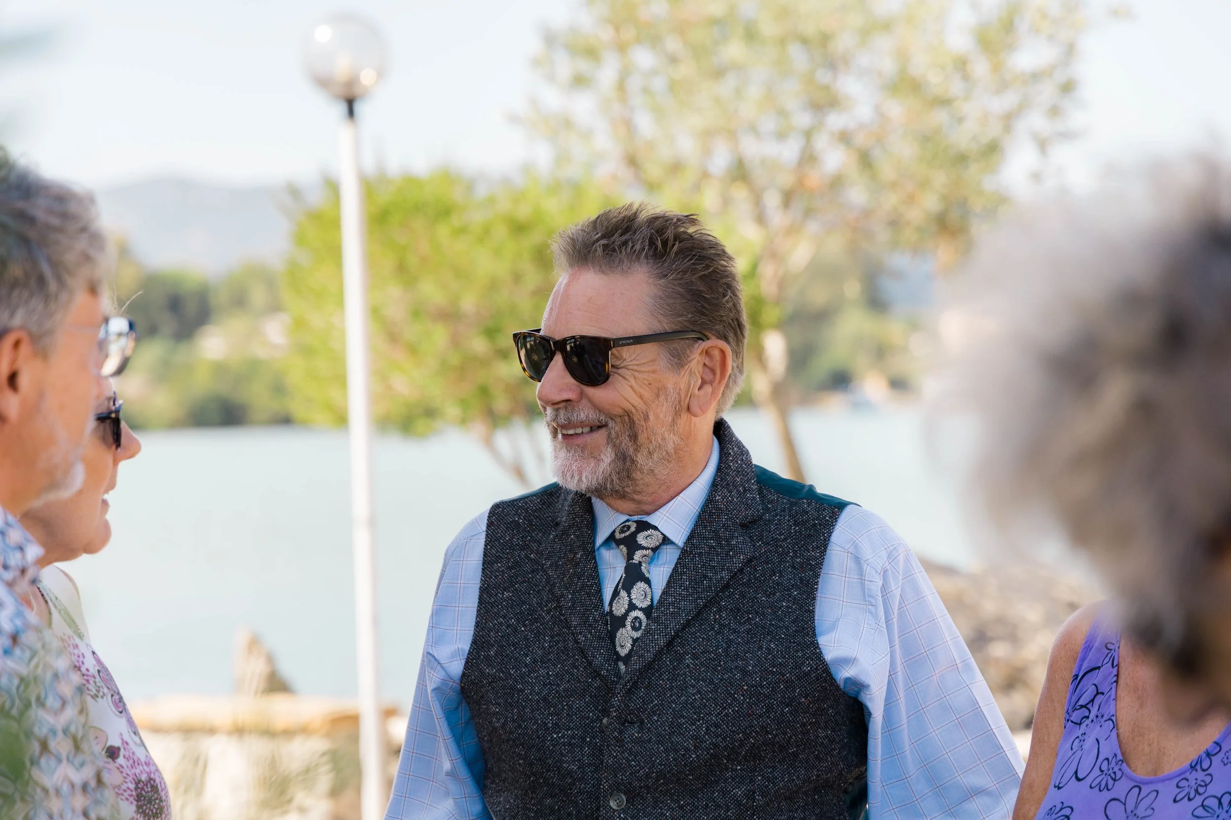 A group of older adults are having a conversation outdoors near a body of water, with trees and a lamp post in the background. The man in the center is wearing sunglasses, a blue checked shirt, a patterned tie, and a dark vest, smiling as he talks to